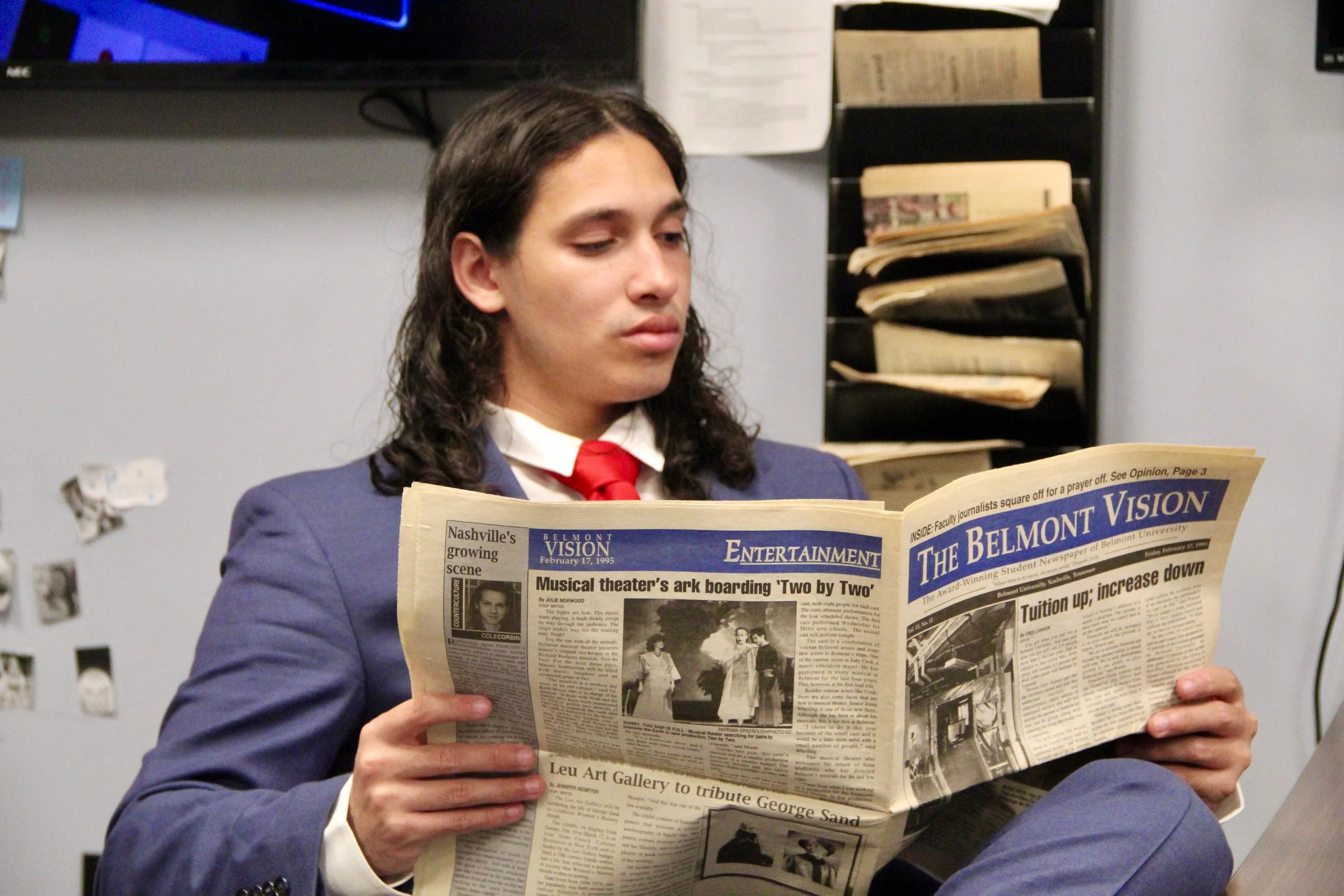 A man with long dark hair and light skin, dressed in a suit and red tie, is reading a newspaper titled 'The Belmont Vision' in an indoor setting with filing shelves and a monitor in the background.