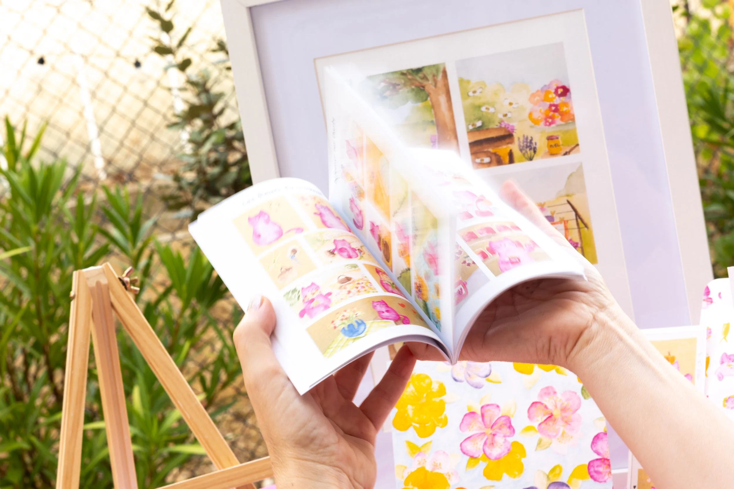 Une personne tient un livre illustré avec des peintures colorées de fleurs et de scènes naturelles, dans un environnement extérieur avec des plantes et un panneau blanc pour exposer l'art.