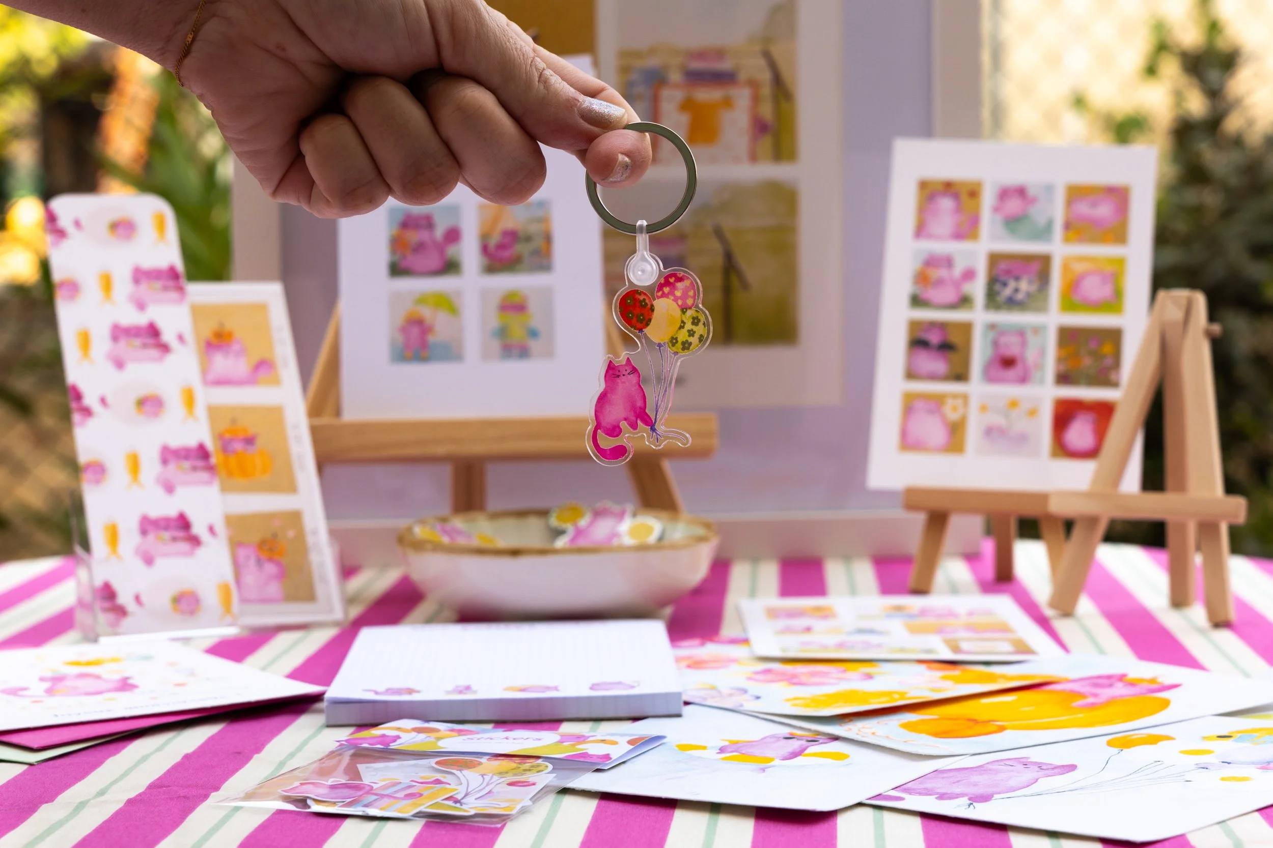 Une main tient un porte-clés avec un pouf en forme de chat et des ballons à motifs, avec un stand de vente d'autocollants et d'illustrations style cartoon en arrière-plan.