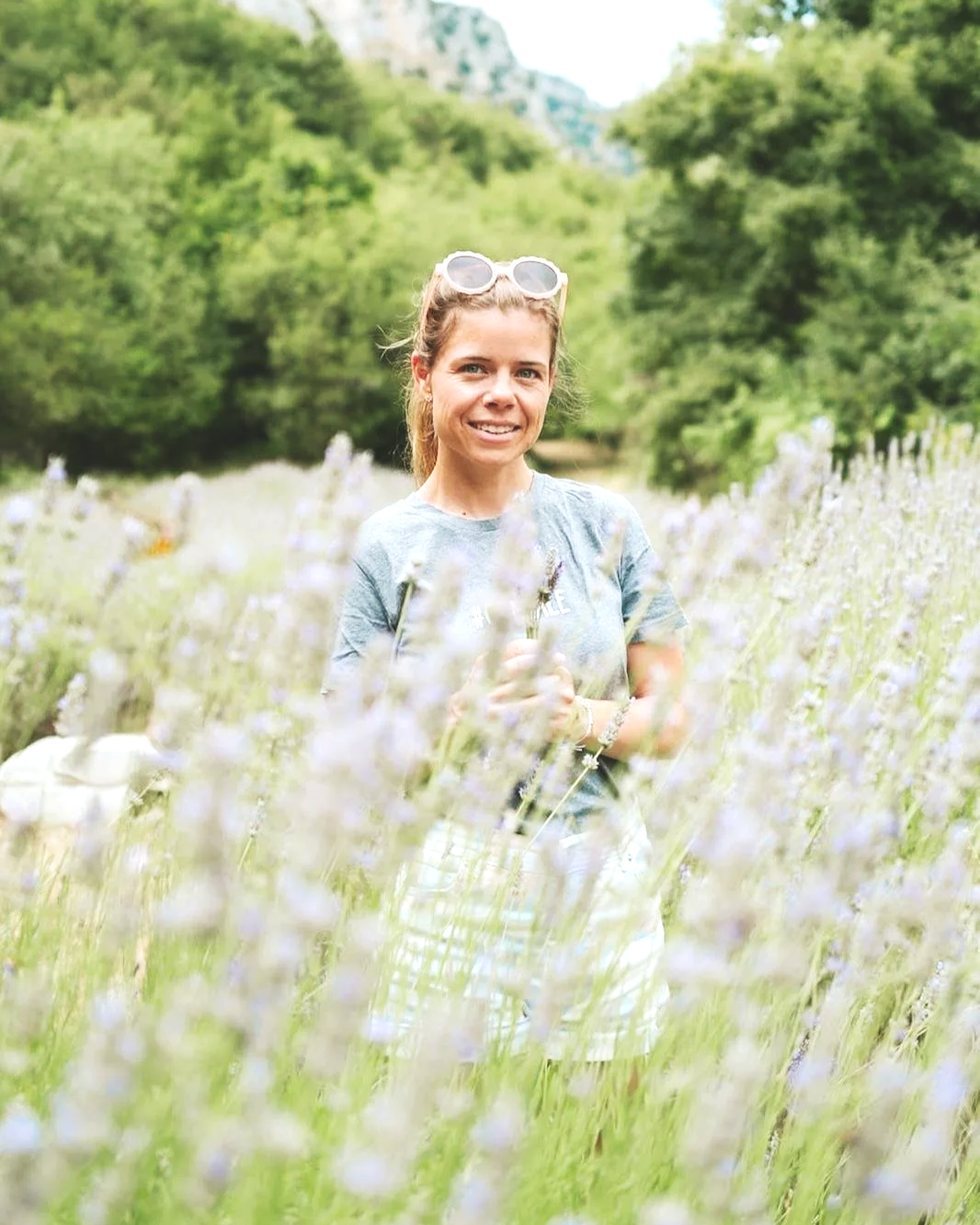 Femme souriante portant des lunettes de soleil, debout dans un champ de lavande, avec des arbres verts en arrière-plan.