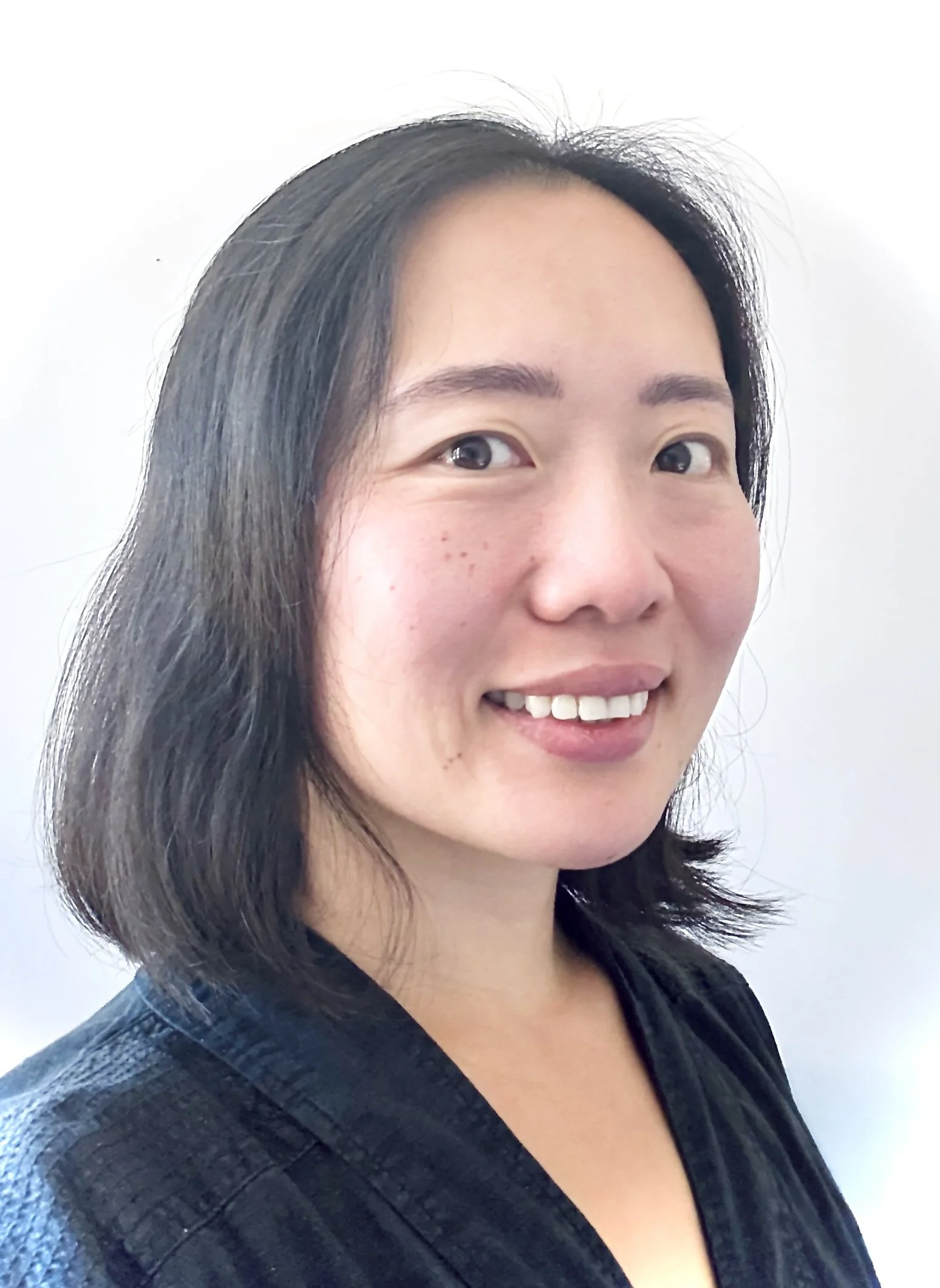 Close-up portrait of a woman with long dark hair, smiling, wearing a black textured top, against a plain white background.