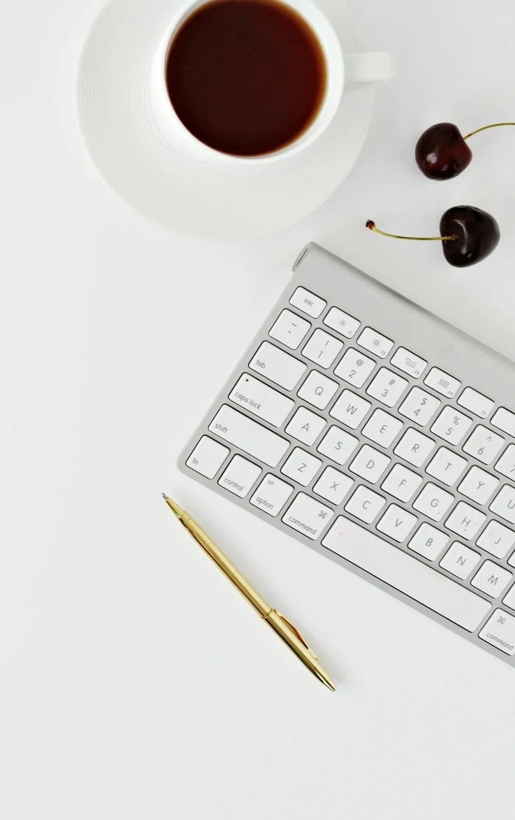 A white computer keyboard, a gold pen, a cup of coffee, and two cherries on a white surface.