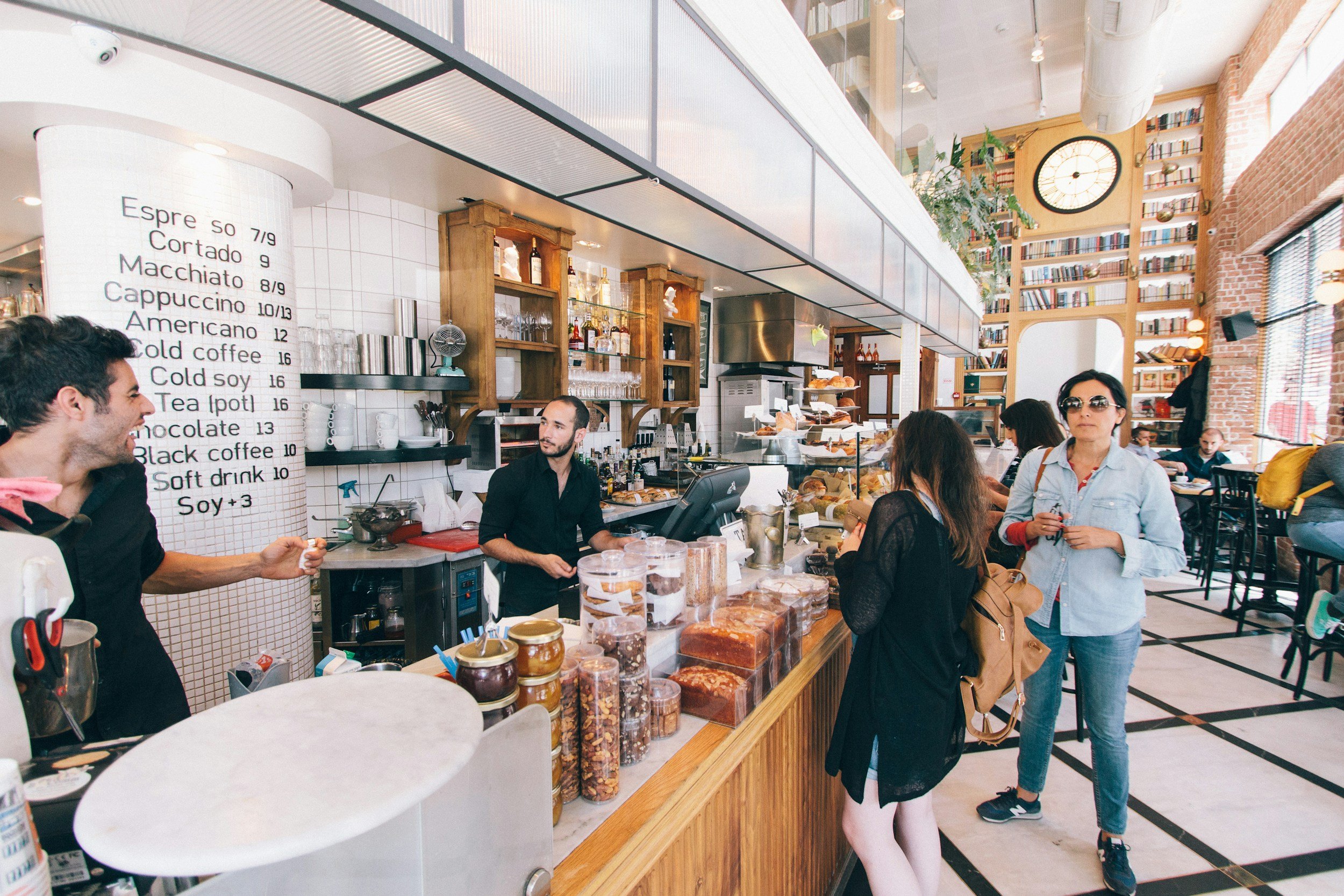 Inside a modern coffee shop with customers standing at the counter. The menu board lists various coffee drinks and prices. Shelves with bottles and glassware are behind the counter. The shop features high ceilings, a large clock on the wall, and a bookshelf-filled upper level. Customers are engaging with baristas and selecting items.
