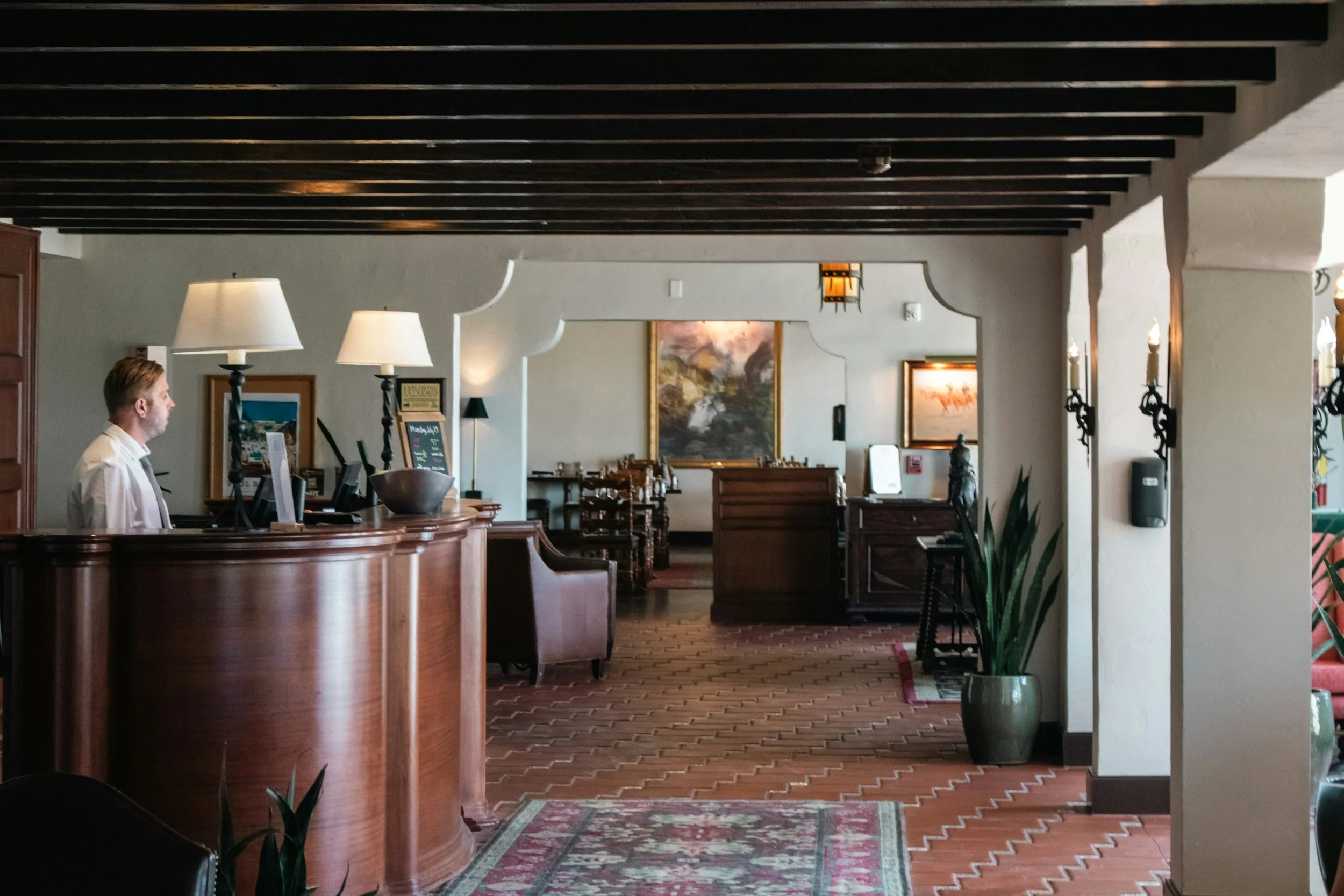 Interior of a restaurant or hotel lobby with a curved wooden reception desk, framed artwork on walls, table lamps, and chairs in the background, and potted plants near a decorative column.