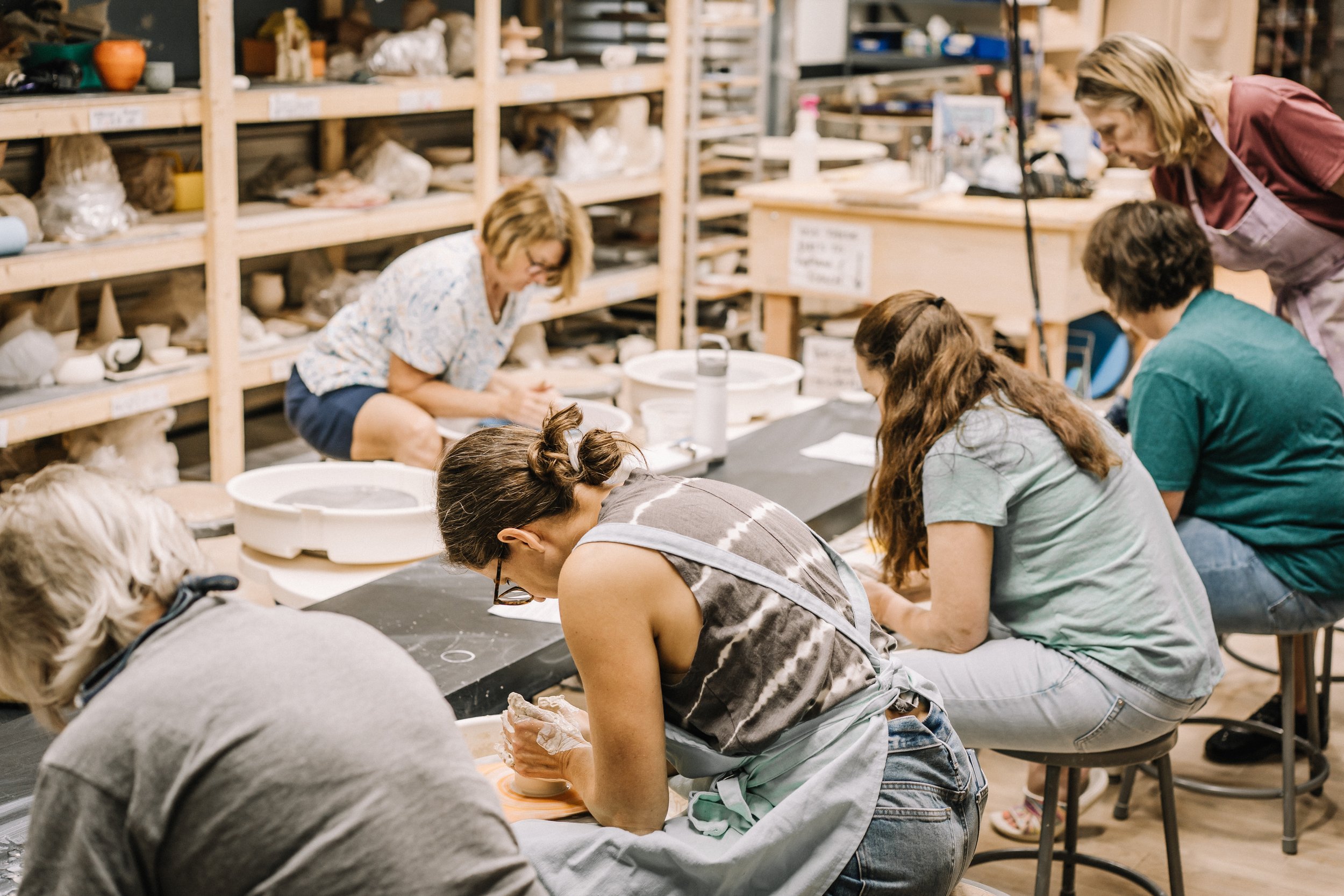 A group of people working on pottery projects in a ceramics studio, with shelves filled with clay and pottery in the background.