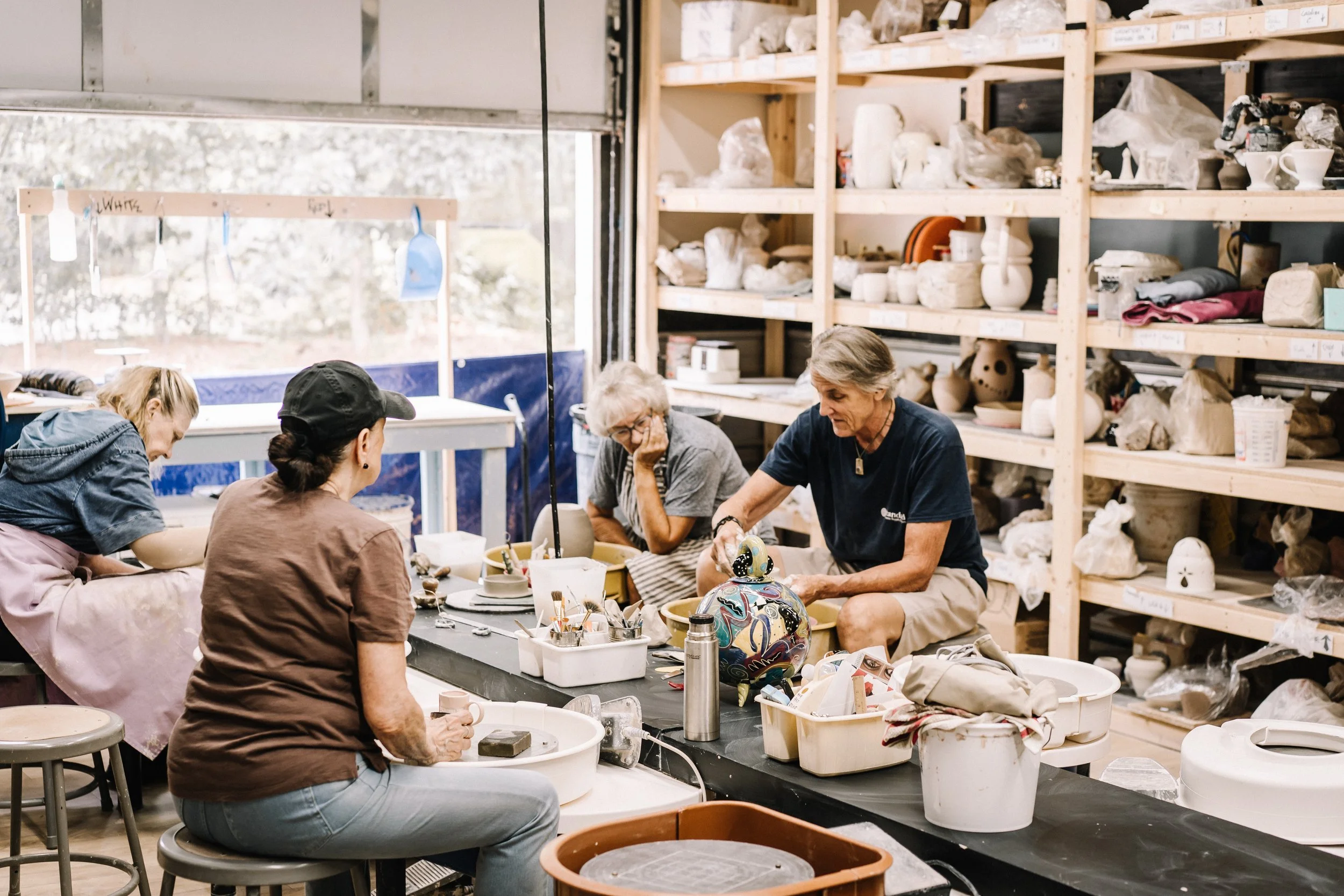 People working on pottery projects in a ceramics studio with shelves of clay and finished pieces in the background.