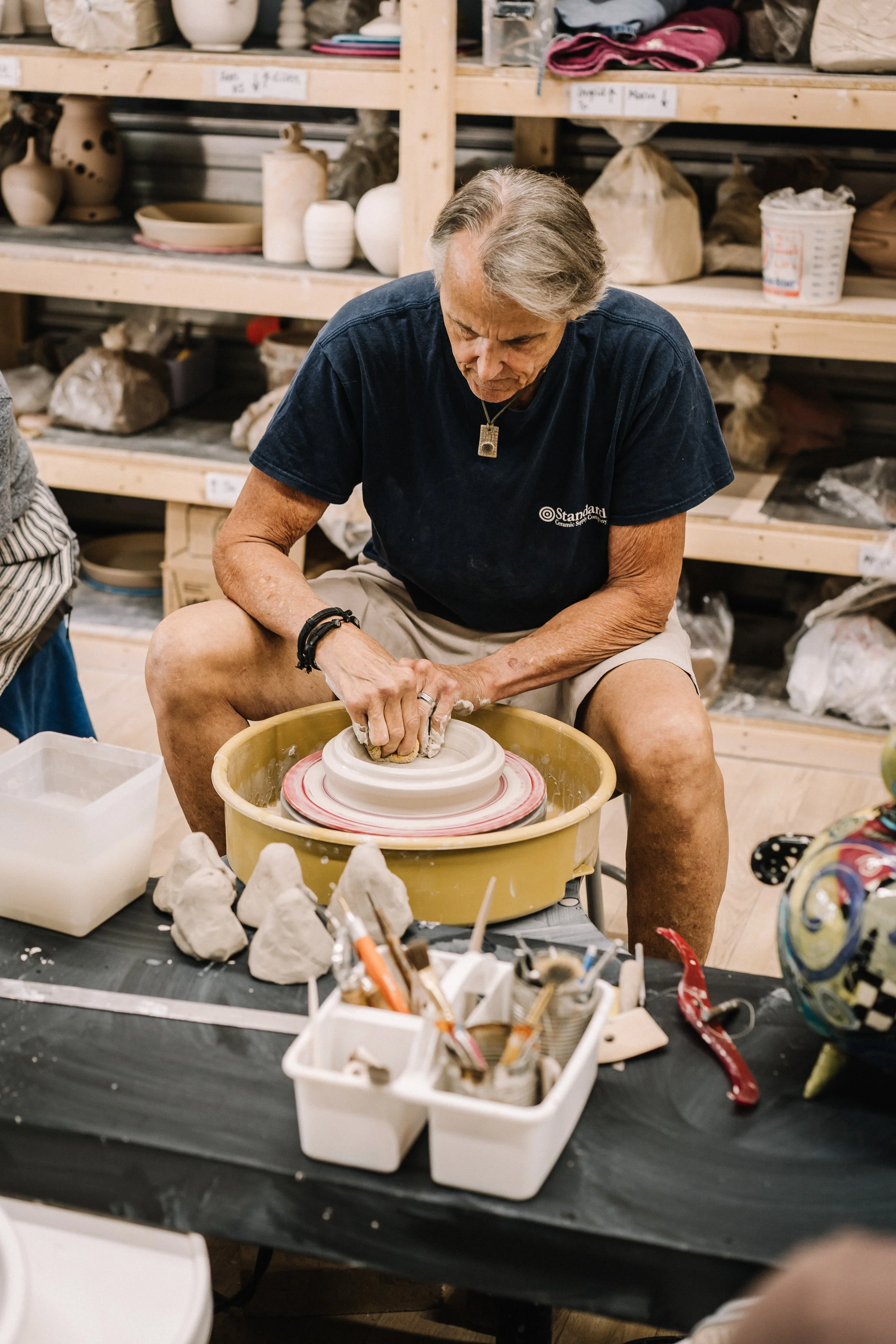 An elderly man working on a pottery wheel in a ceramics studio surrounded by shelves filled with ceramic pieces and tools.