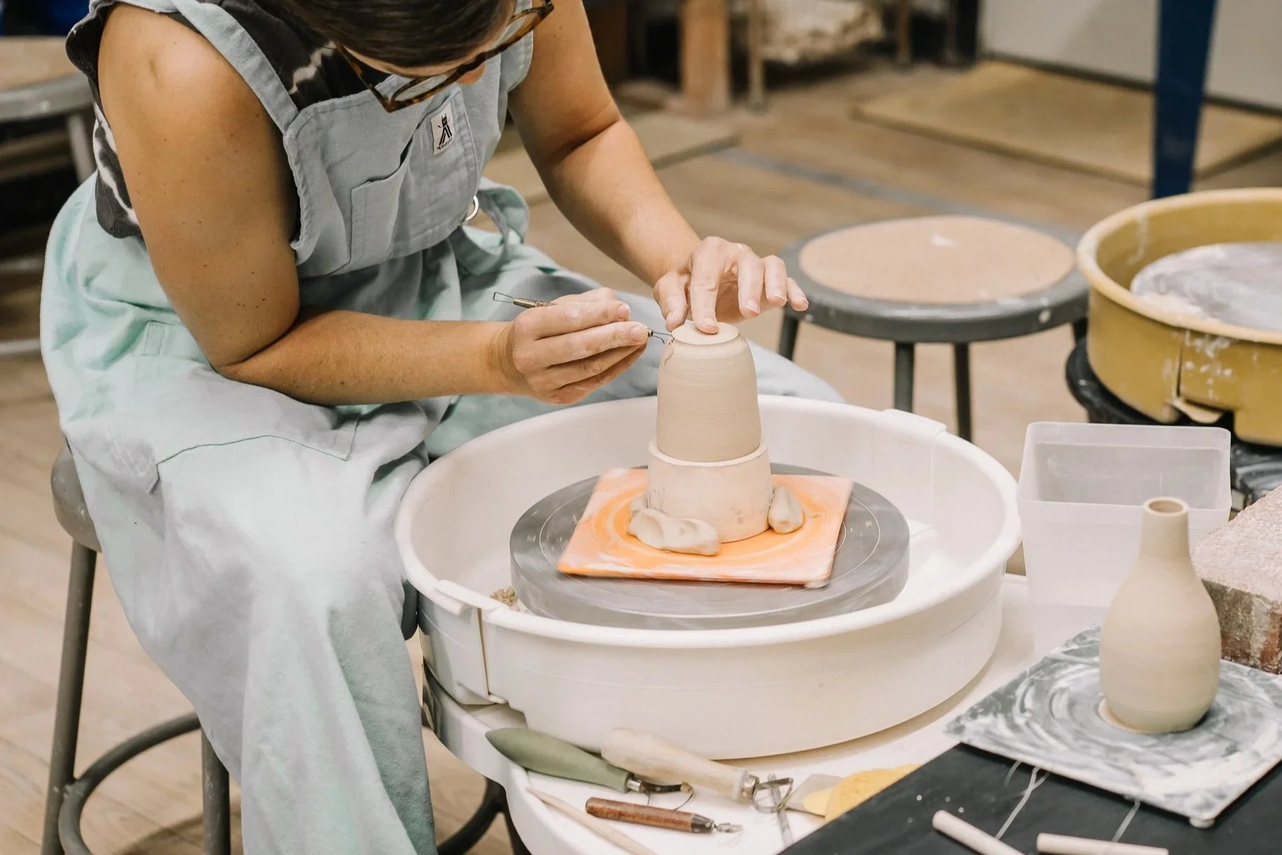 A woman sitting at a pottery wheel, shaping a tall ceramic vase, with pottery tools and supplies around her in a workshop.