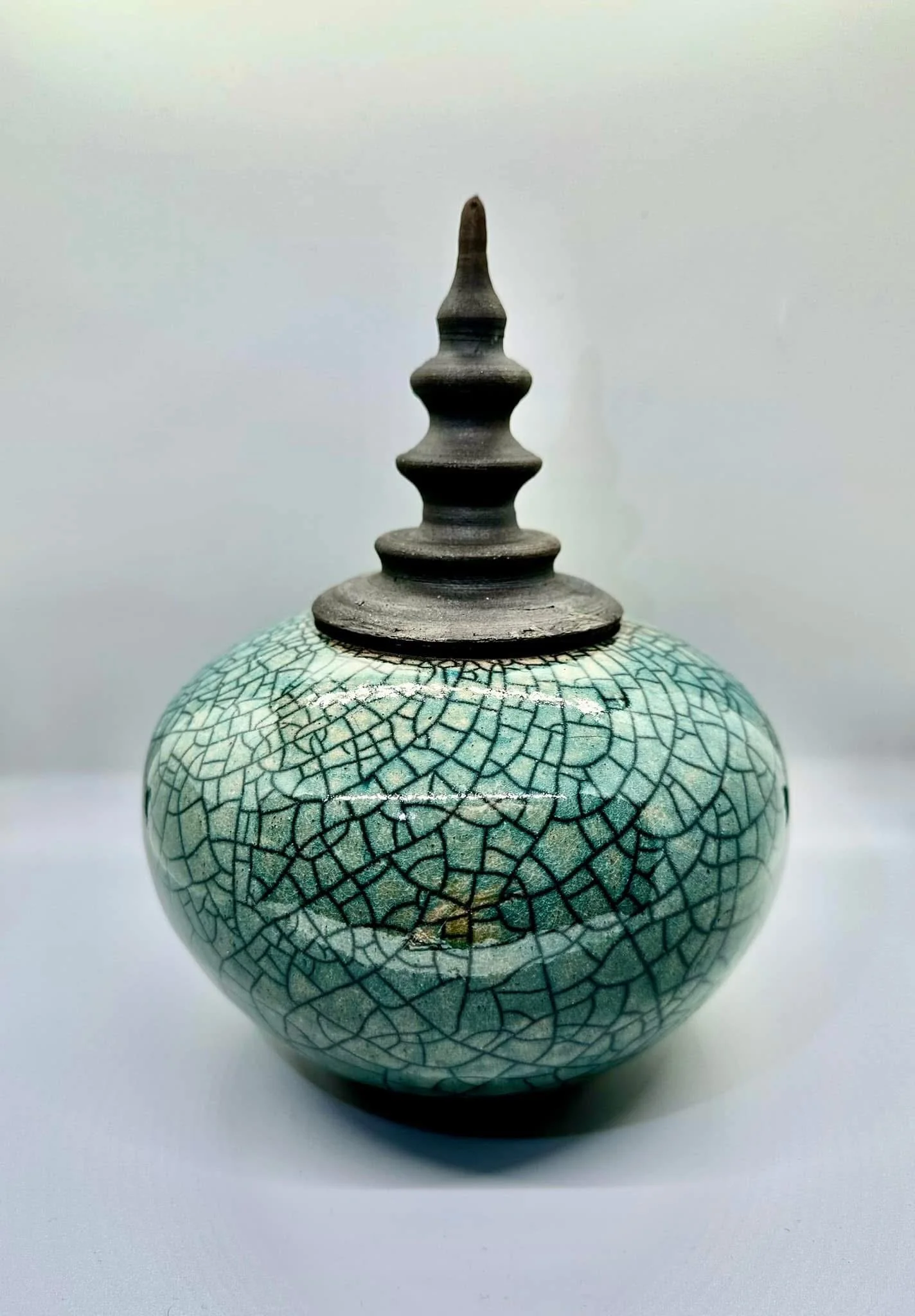 Round ceramic vase with a cracked glaze pattern in shades of green and black, featuring a metal or dark wood lid with a pointed finial on top.