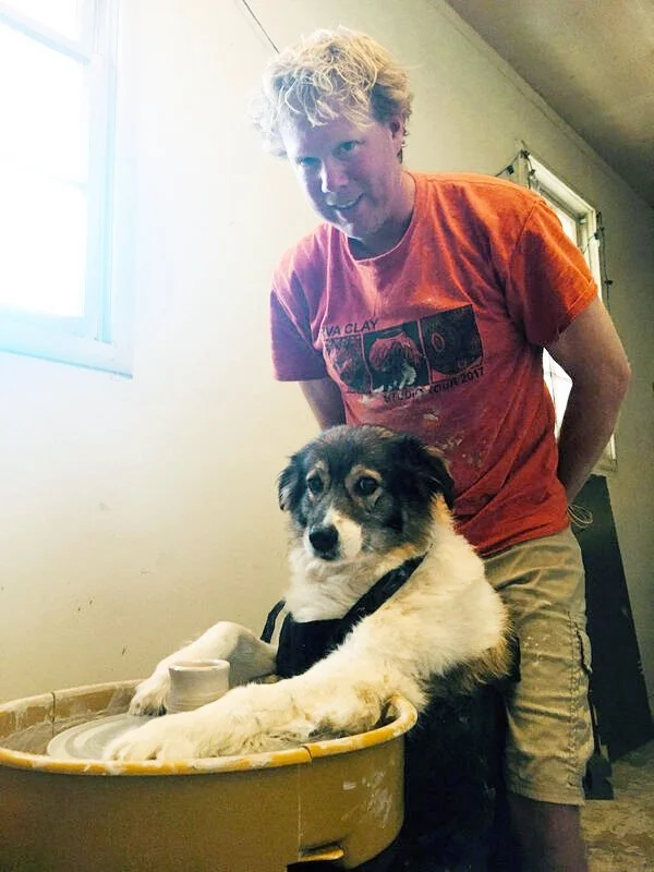A young man with curly blonde hair leaning over a young dog in a yellow plastic tub. The dog is lying down with one paw resting on the edge of the tub, wearing a black collar and looking at the camera. The setting appears to be indoors near a window, with a wall in the background.