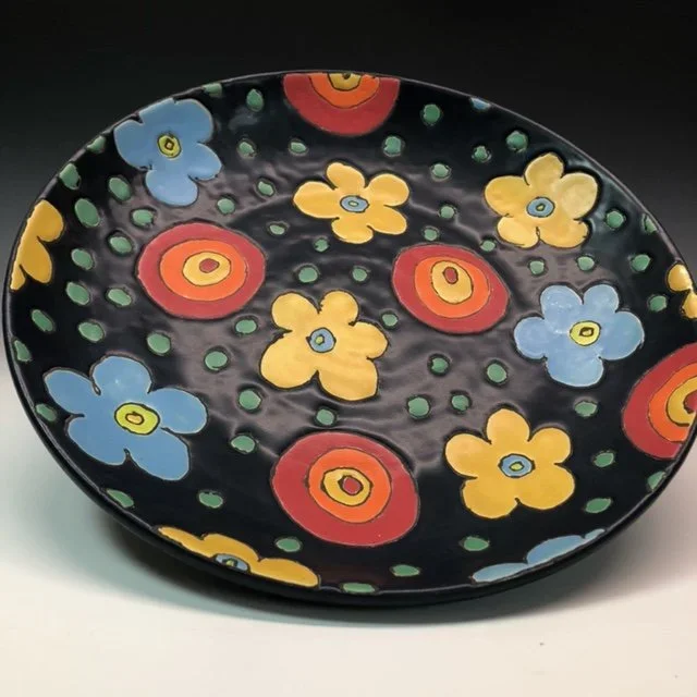 Decorative black ceramic plate with colorful floral pattern in yellow, blue, and red with green dots.