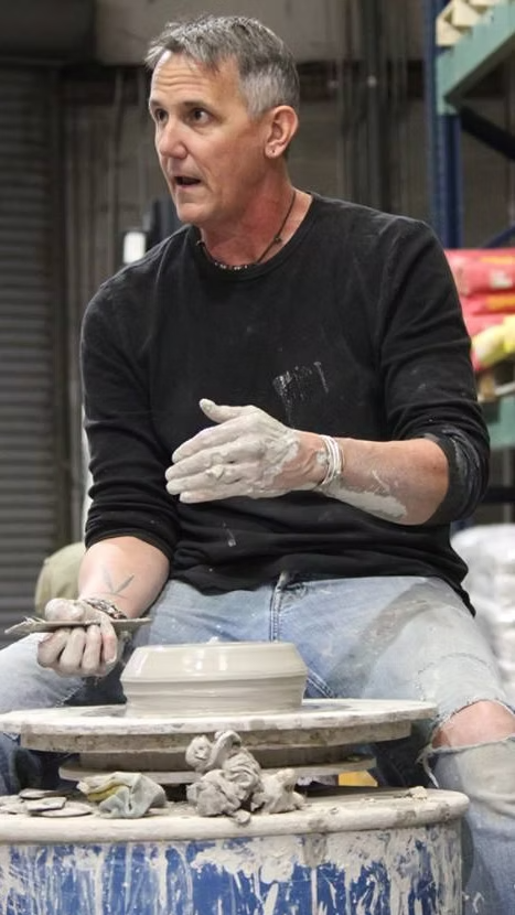 A man with short gray hair, wearing a black long-sleeve shirt, is working on a pottery wheel in a studio. He has clay on his hands and jeans with ripped knees, and he appears to be explaining or teaching something.