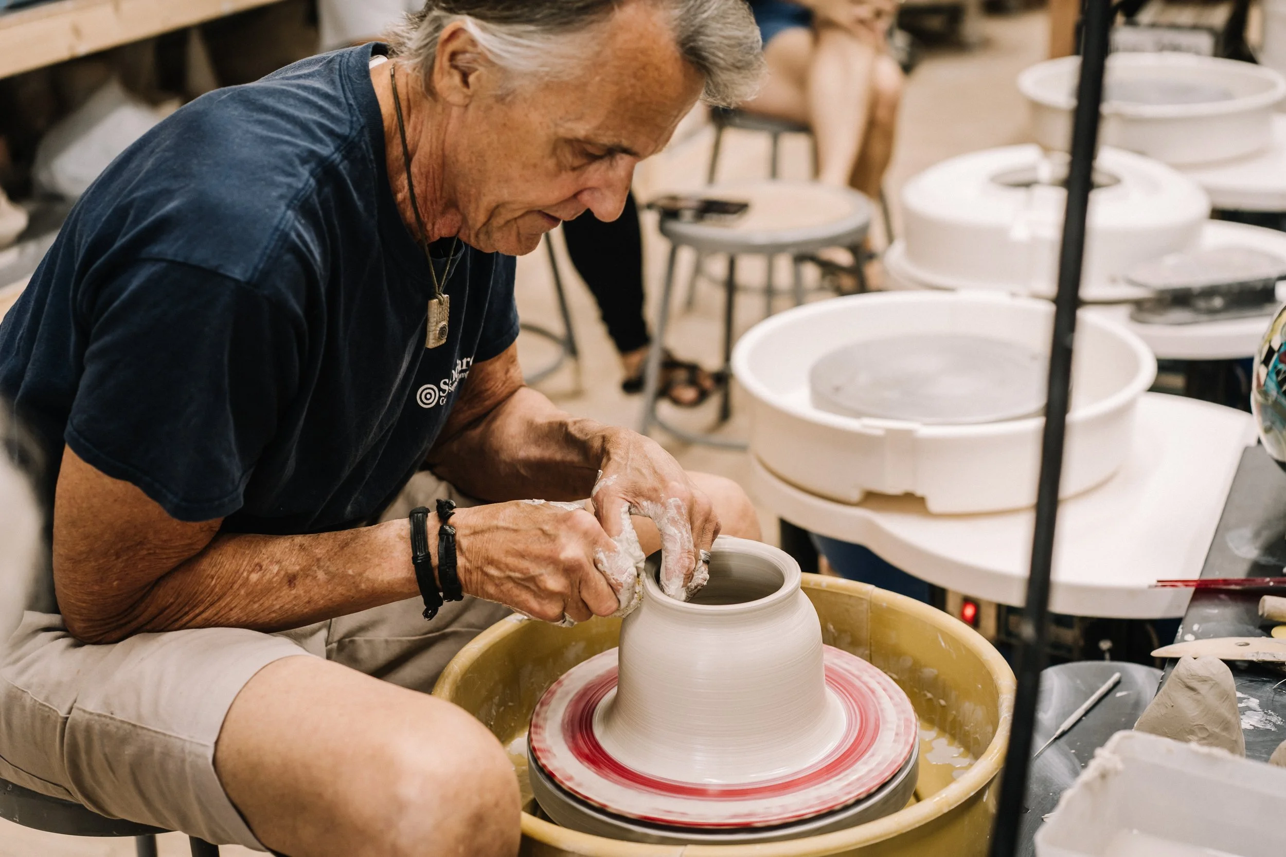 An elderly man working on a pottery wheel shaping a ceramic piece in a studio with shelves of supplies and white buckets in the background.
