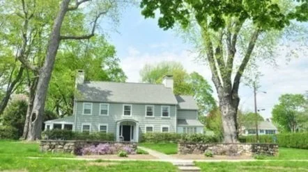 A large gray house with multiple stories, surrounded by tall trees and green grass, with a stone wall in front and a sidewalk leading to the entrance.