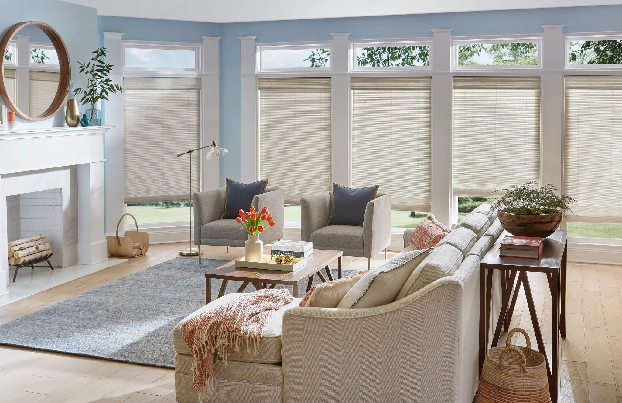 Cream cellular shades on large living room windows in a bright blue and neutral living room with a sofa and two accent chairs