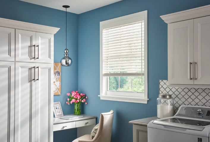White faux wood blinds on a window in a blue home office with a white desk and pendant light