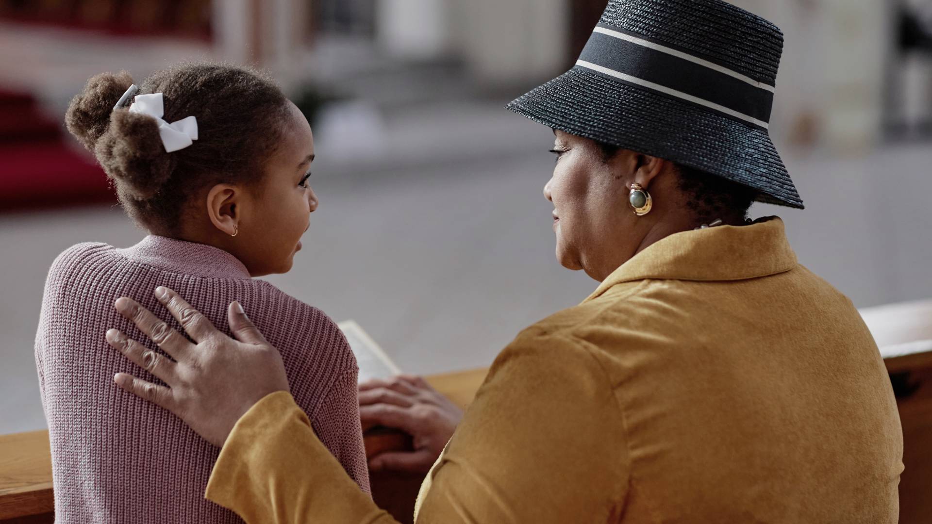 An elderly woman with a black straw hat and gold earrings is embracing and talking to a young girl with hair in a bun, white ribbons, and a pink sweater.