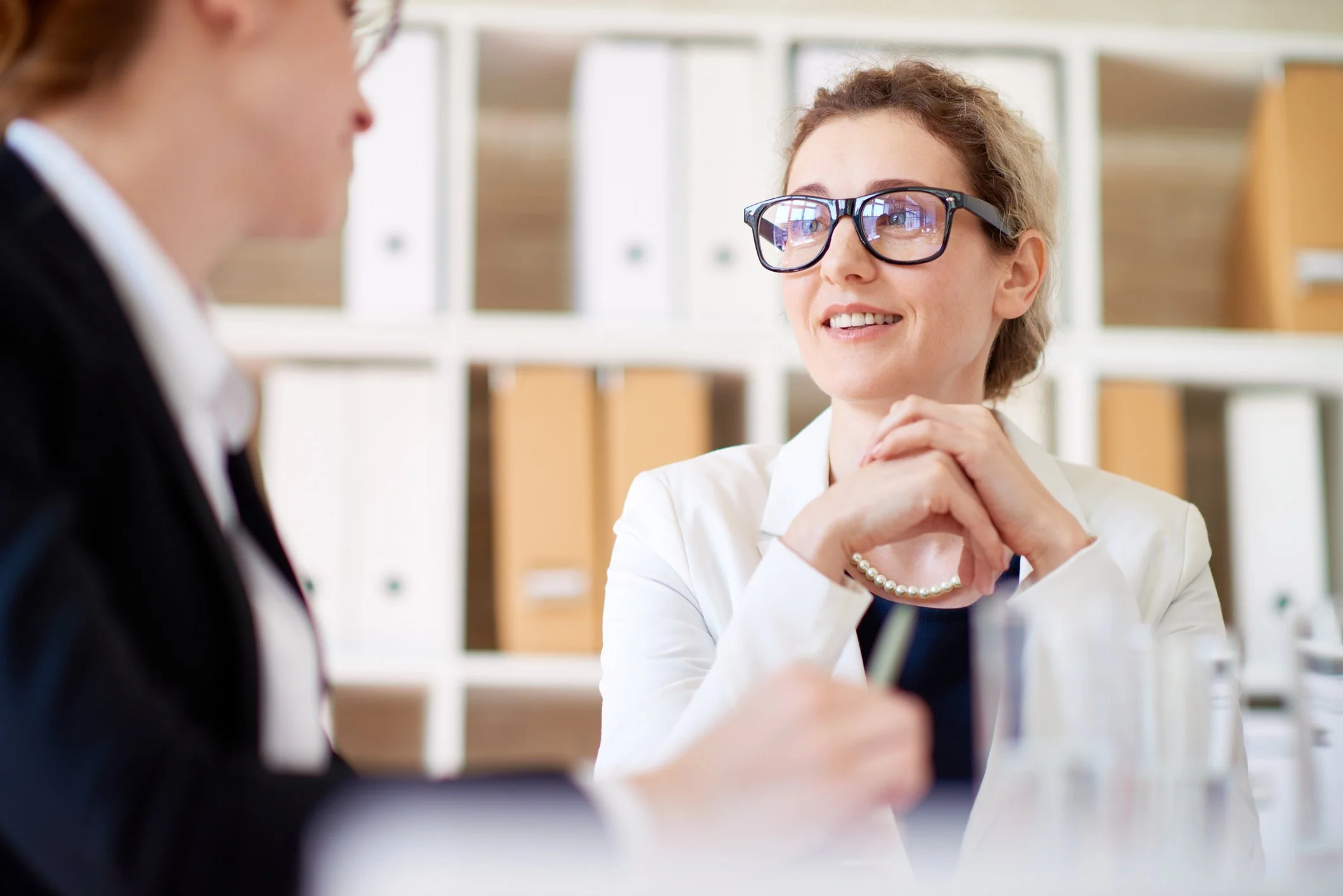 A smiling businesswoman wearing thick black glasses and a white blazer listening intently to a colleague during a professional consultation.