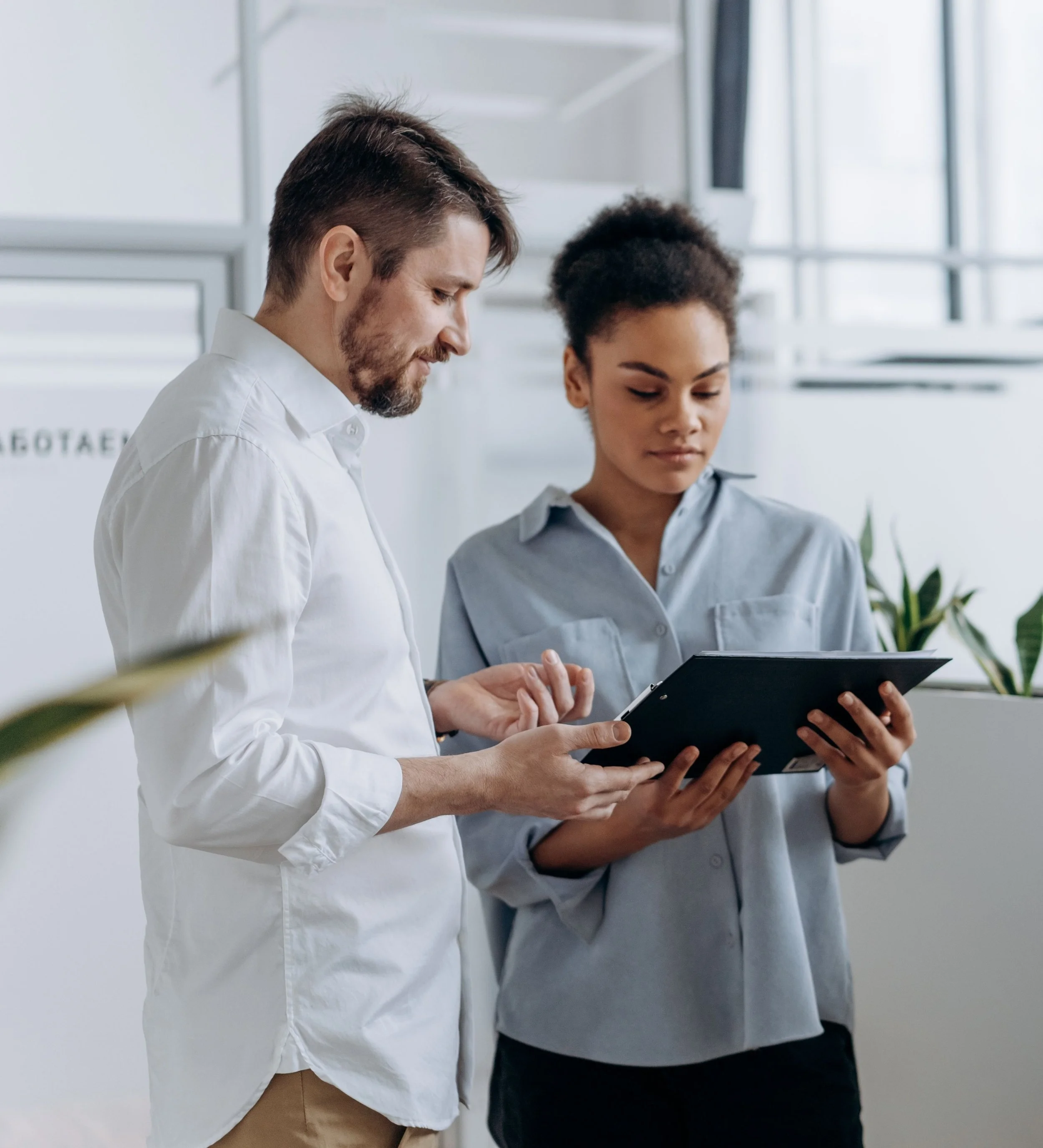 A male and female business professional standing together in a bright, modern office while reviewing data on a black clipboard.
