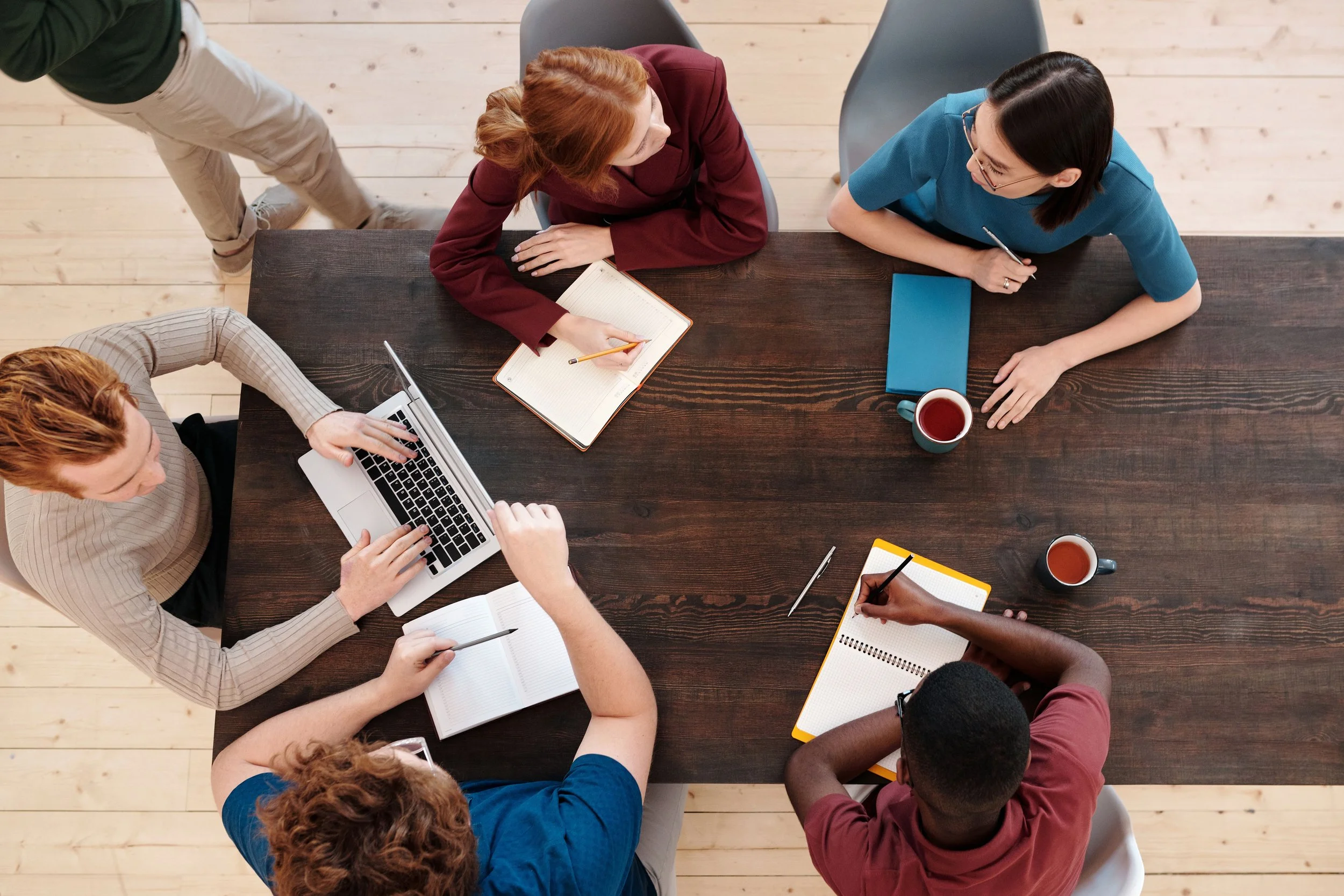 Overhead view of a diverse business team sitting around a conference table, collaborating, taking notes, and typing on a laptop during a workspace meeting.