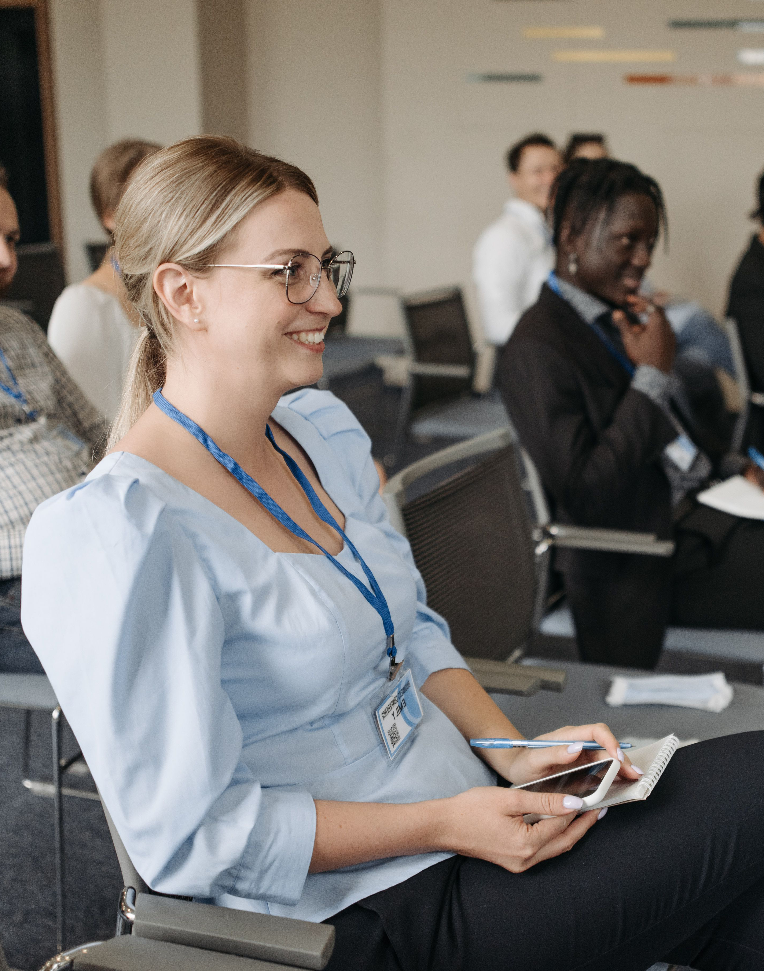 Side angle view of a smiling blonde businesswoman wearing glasses and a blue lanyard name tag, holding a smartphone and pen while sitting in an audience at a professional corporate seminar.