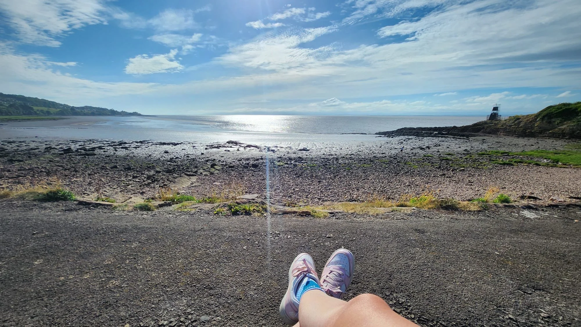 Person in pink and blue sneakers sitting on a concrete surface staring at the ocean, with a rocky shoreline, grassy hill, and a lighthouse in the distance under a partly cloudy sky.