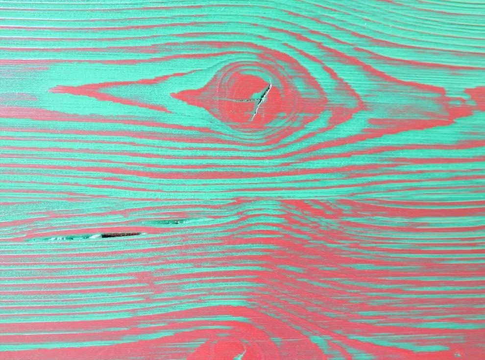 Close-up of a weathered wooden surface with pink and teal stain patches, visible wood grain, and cracks.