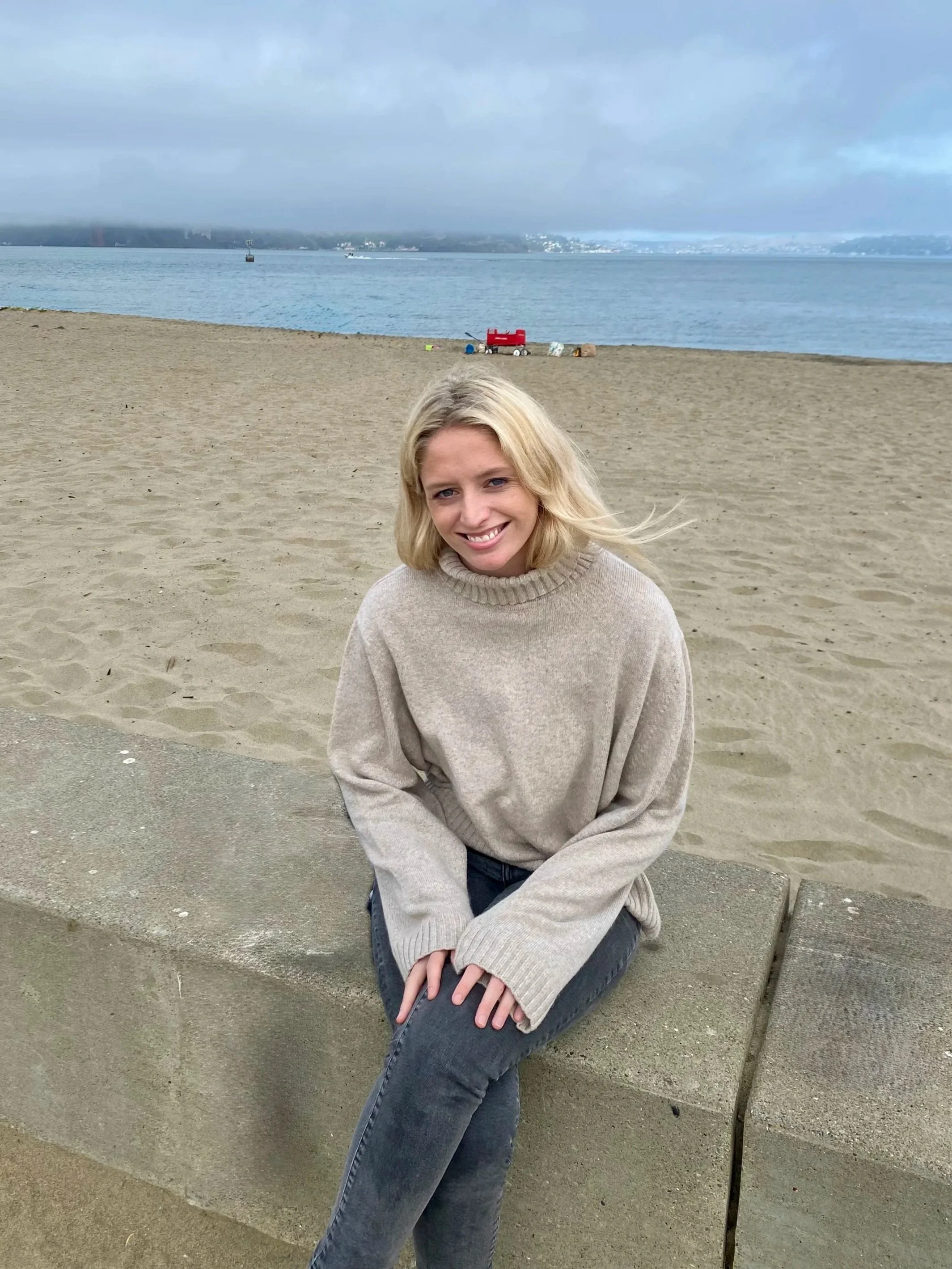 A young woman with blonde hair smiling while sitting on a concrete bench at the beach. She is wearing a beige sweater and dark jeans. The background shows sand, water, boats, and a cloudy sky.