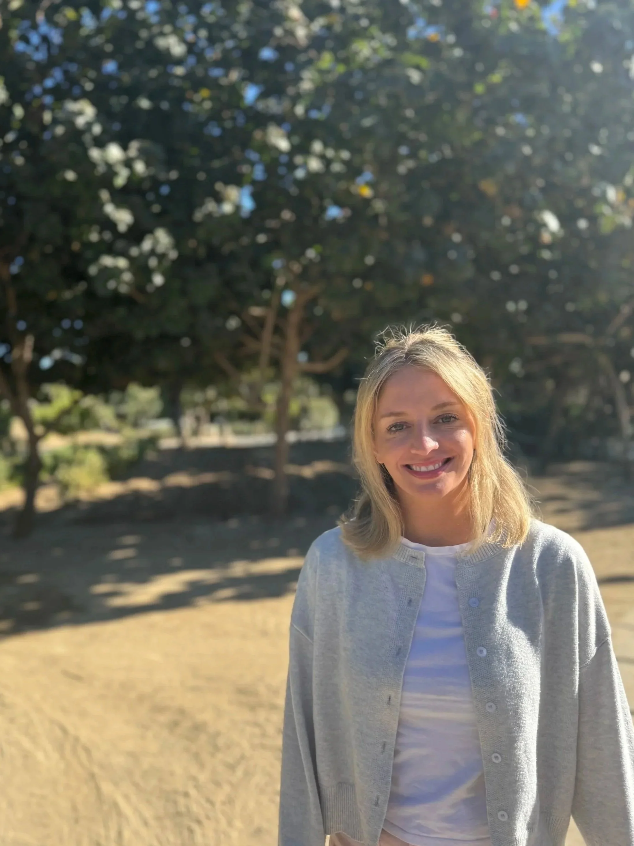 A woman with blonde hair smiling outdoors with trees and a grassy area in the background.