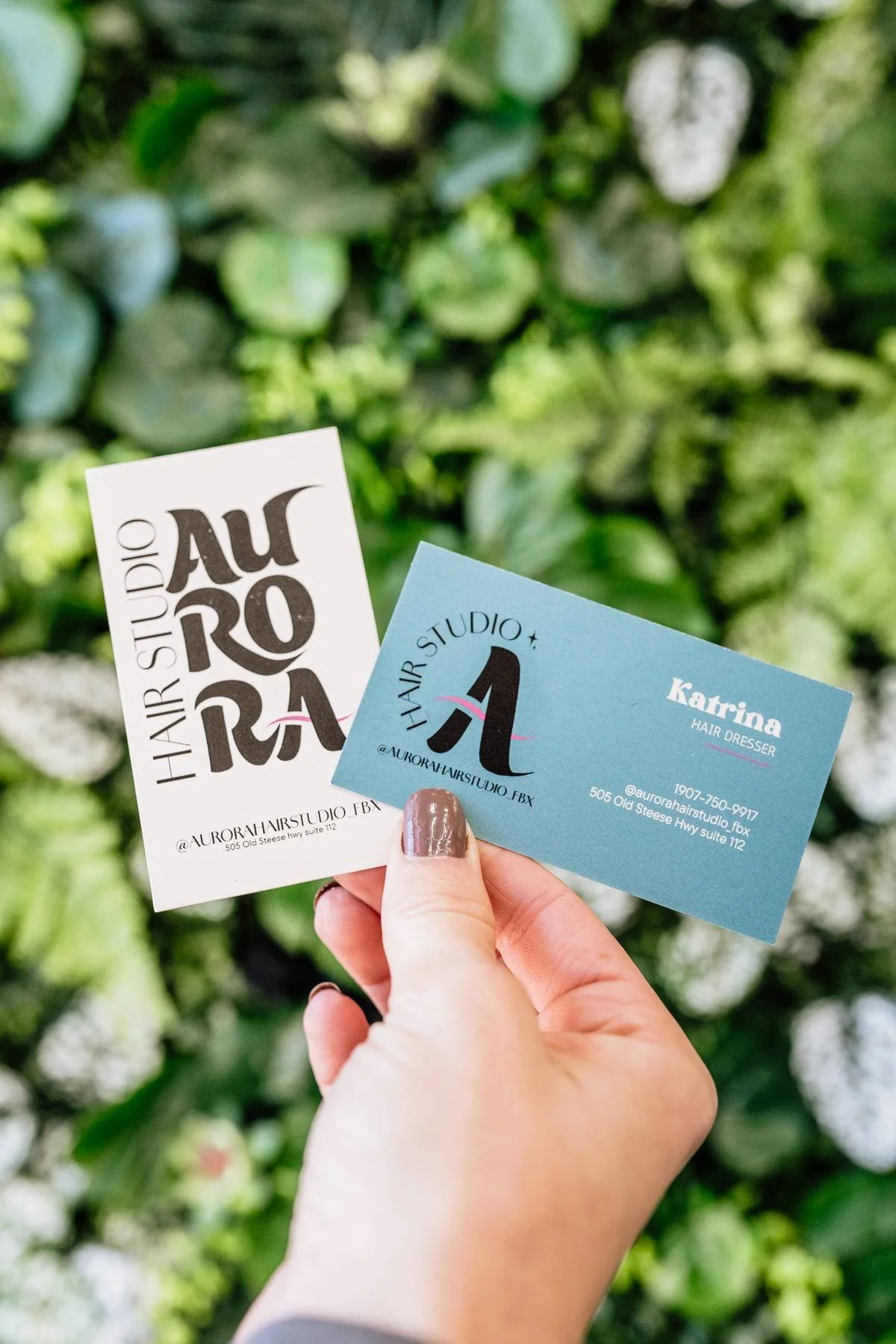 Hand holding two business cards for Aurora Hair Studio against a blurred green foliage background.