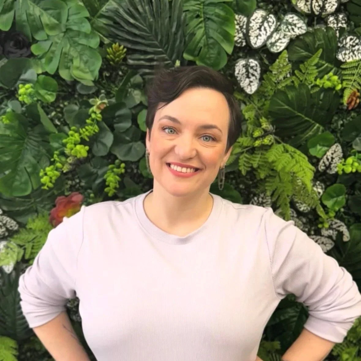 Woman with short dark hair smiling in front of a lush green leafy wall.