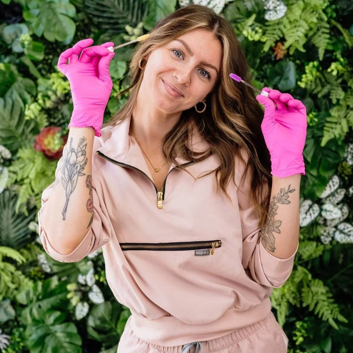A woman with tattoos, wearing pink gloves, holding two syringes, smiling, standing in front of a green leafy background.