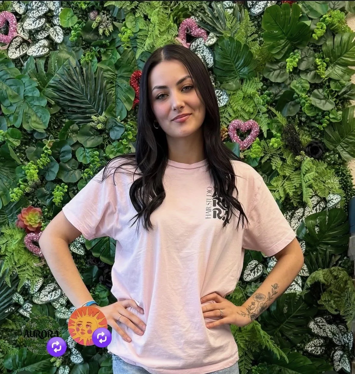 A woman with long dark hair wearing a light pink T-shirt, standing in front of a green leafy background with pink heart-shaped decorations.