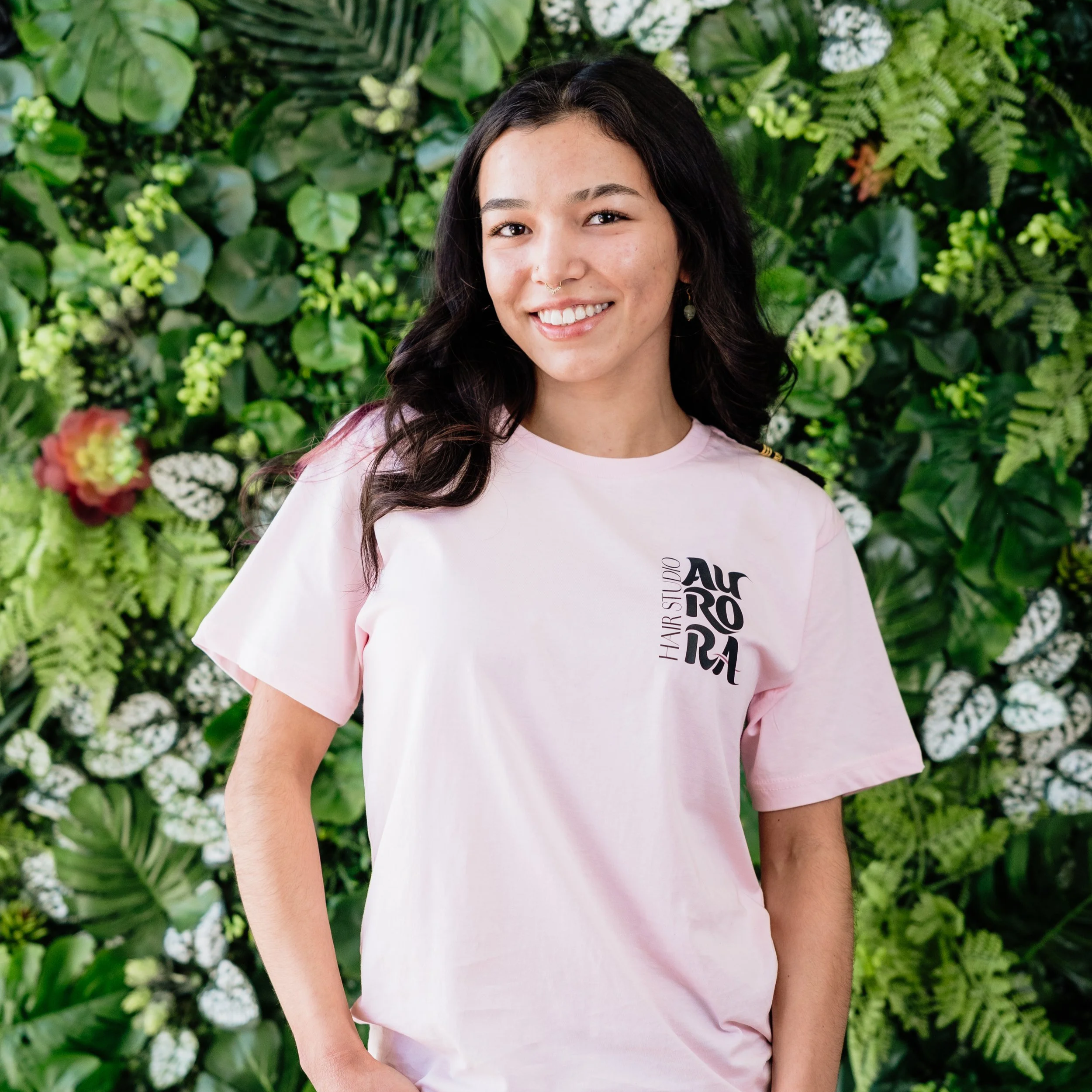 A young woman with long dark hair smiling, wearing a pink T-shirt, standing in front of a lush green wall of various plants.