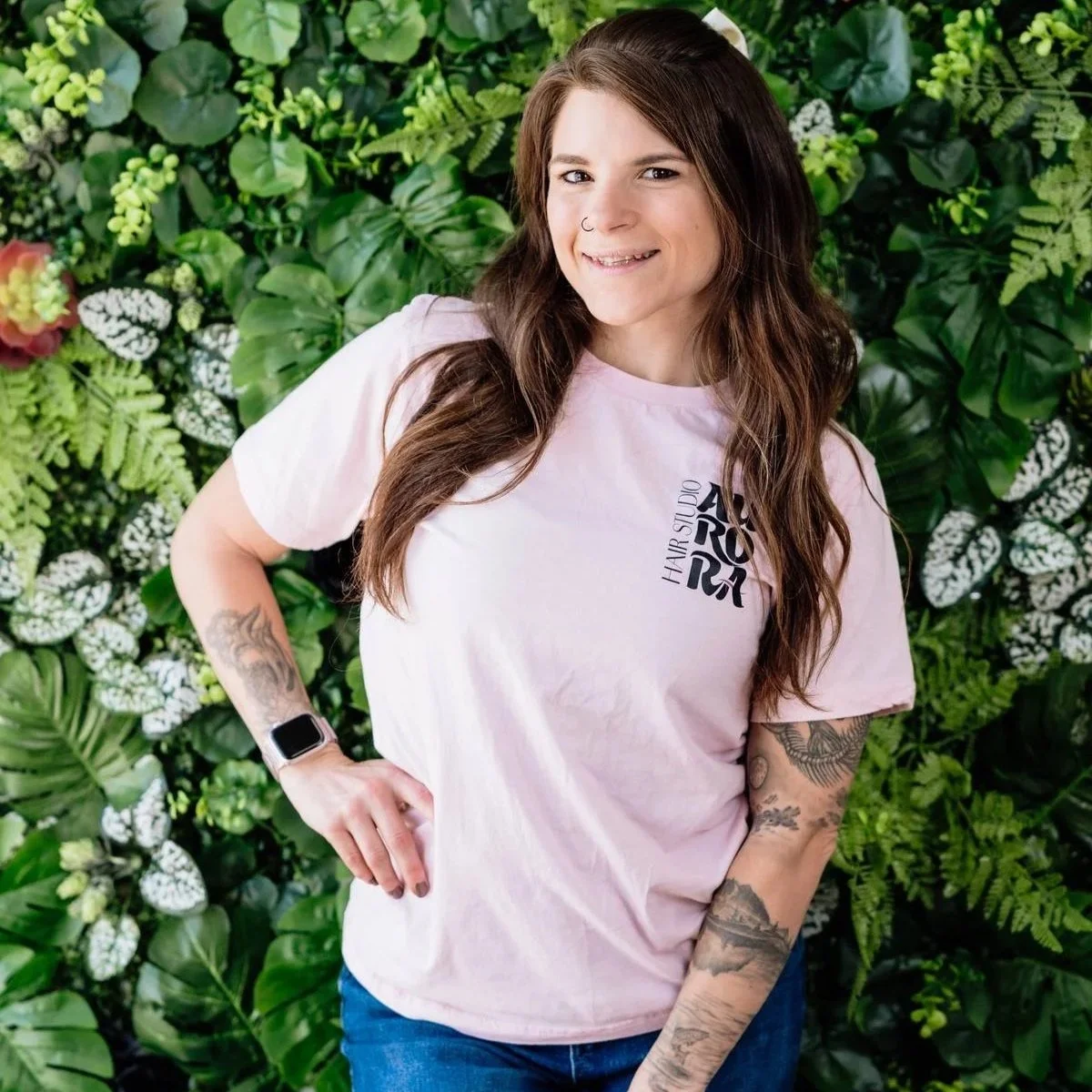 A young woman with long brown hair, tattoos on her arms, and a nose ring, posing in front of a leafy green background. She is wearing a pink T-shirt with black text and blue jeans.
