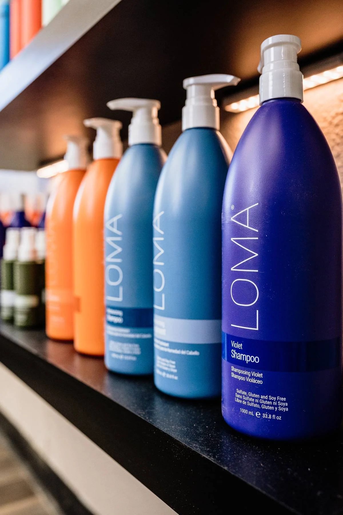 A row of colorful bottles of Loma brand shampoo on a black shelf, with the ones in focus being purple and blue, labeled as violet shampoo and shampoo respectively.