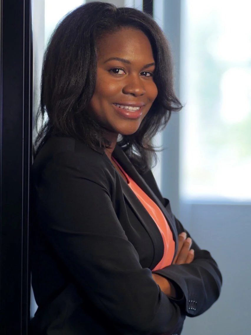 Professional headshot of an African American woman with shoulder-length dark hair, wearing a black blazer over an orange top, standing next to a window with her arms crossed and smiling at the camera. African American therapist.