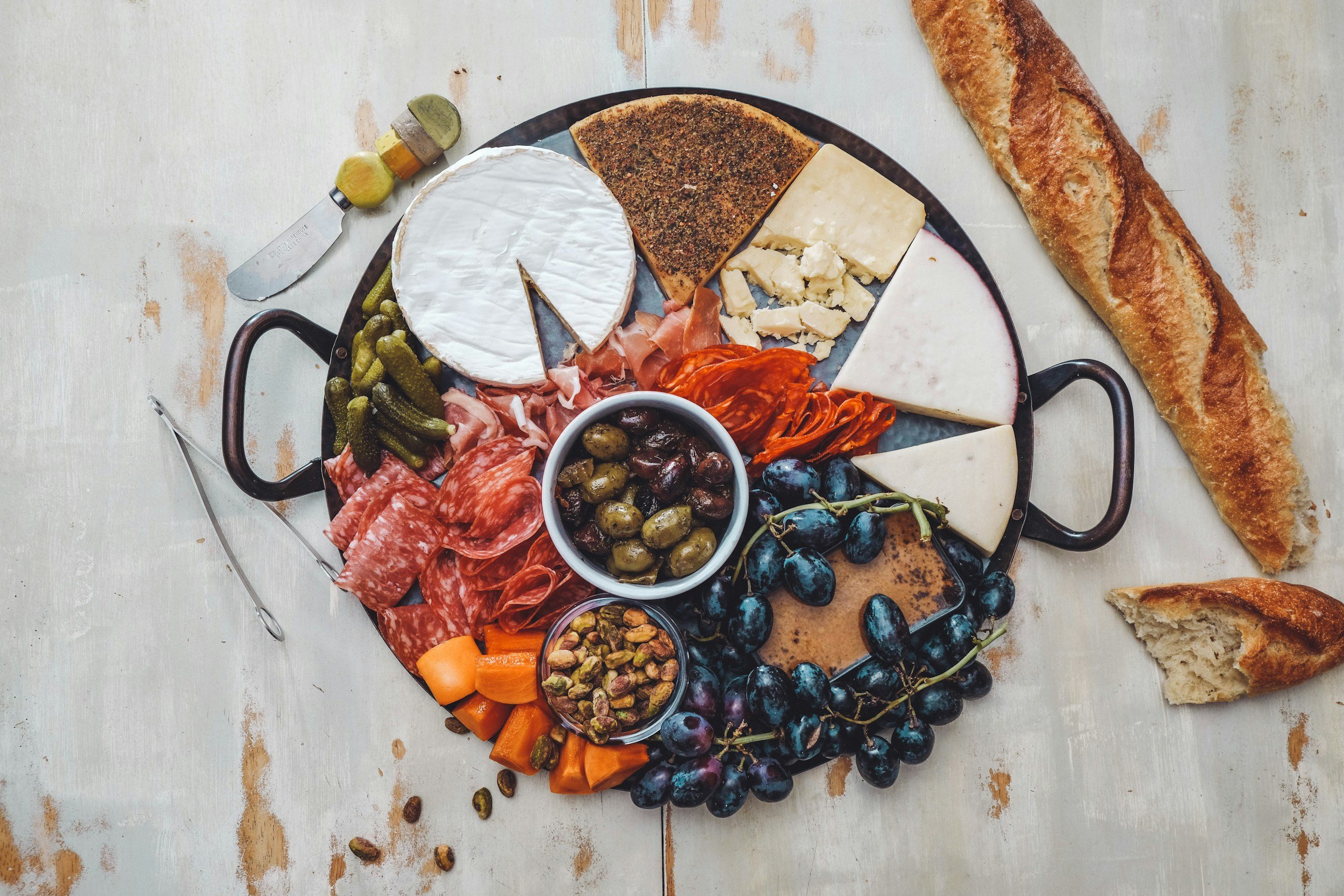 A cheese and charcuterie board with various cheeses, meats, grapes, pickles, nuts, and bread on a round tray, with additional bread on the side.