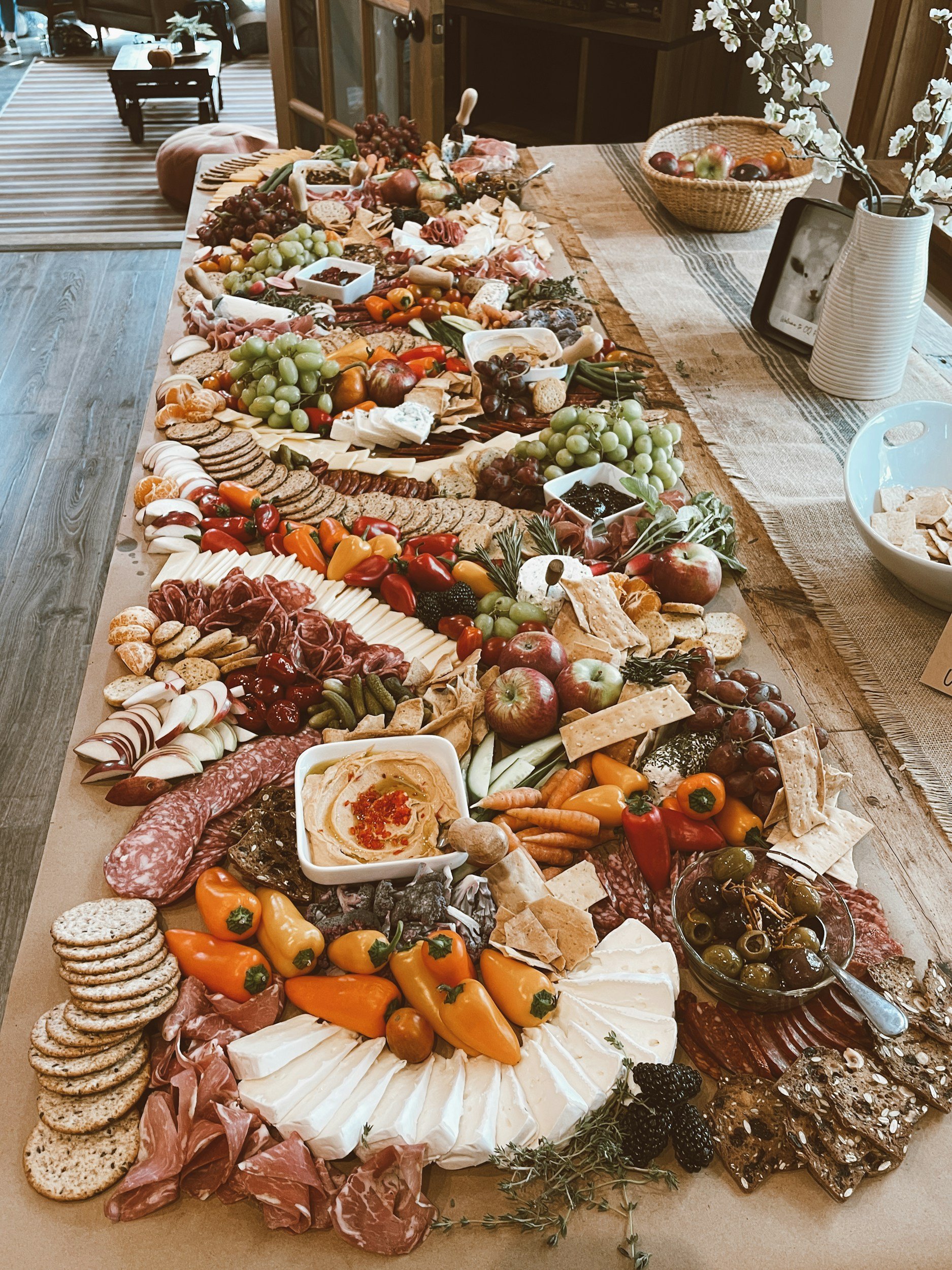 A large charcuterie board with an assortment of cheeses, cured meats, crackers, fruits, vegetables, spreads, and olives, displayed on a table in a cozy indoor setting.