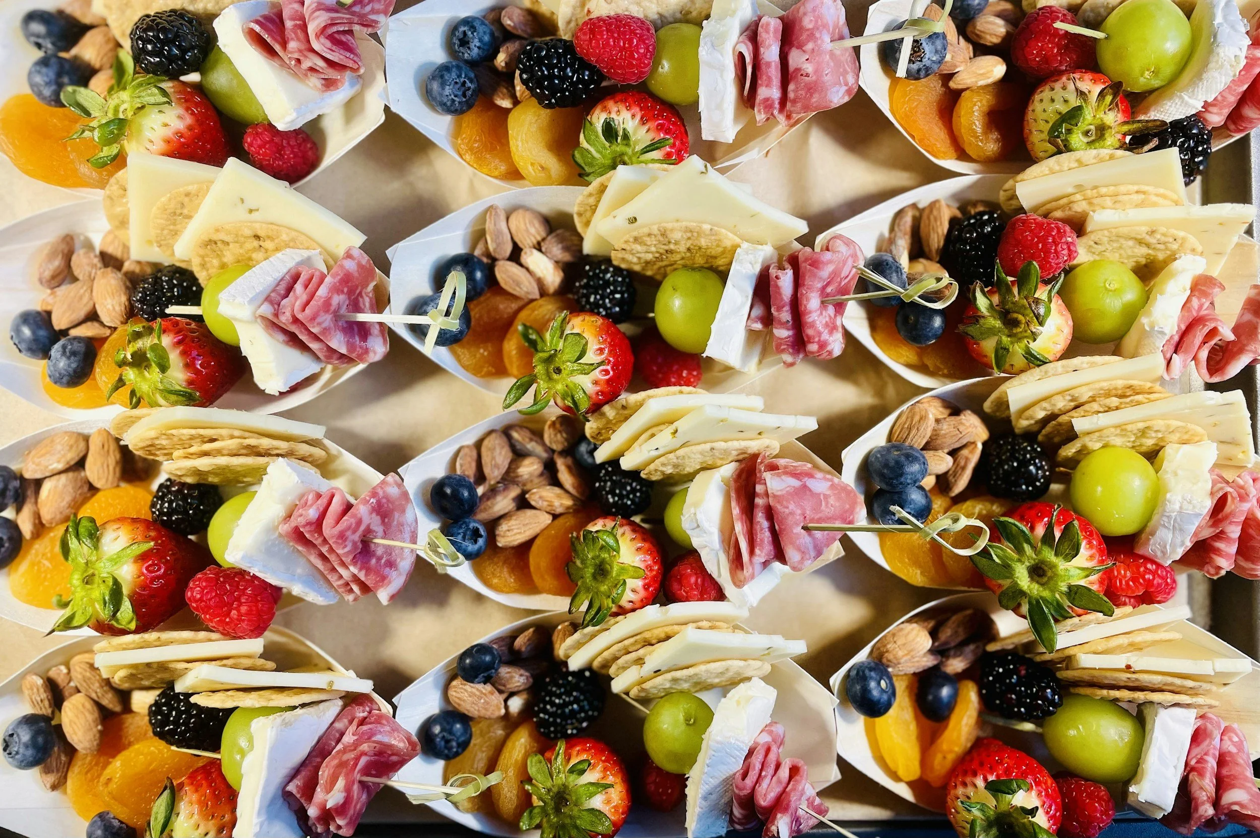Assorted fruit and cheese appetizers with strawberries, blueberries, blackberries, green grapes, mandarin oranges, cheese slices, almonds, and slices of cured meat, arranged in small servings.