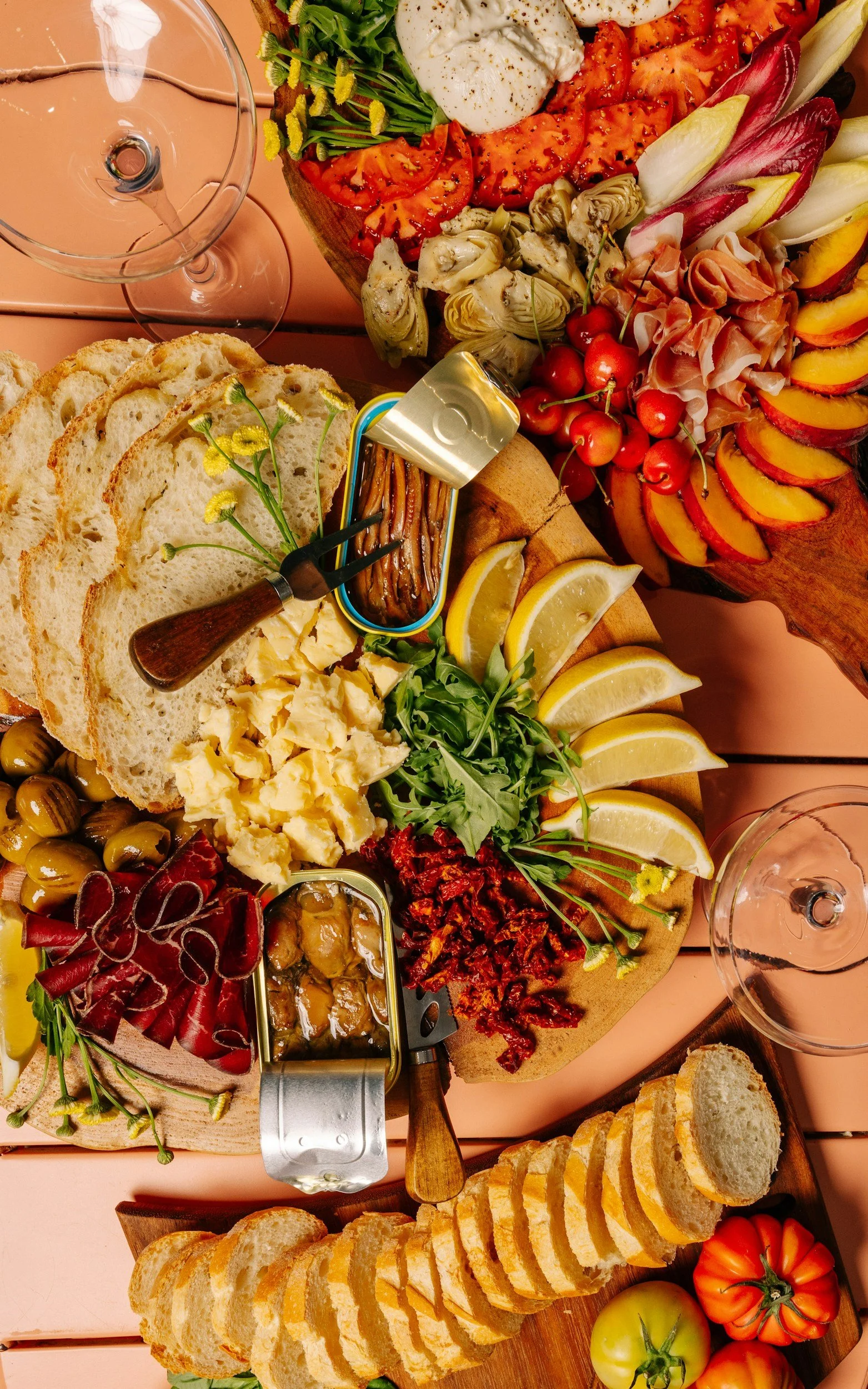 A cheese and charcuterie board with sliced bread, cheeses, olives, cherry tomatoes, sun-dried tomatoes, sliced peaches, and various meats arranged on wooden platters, with wine glasses nearby.
