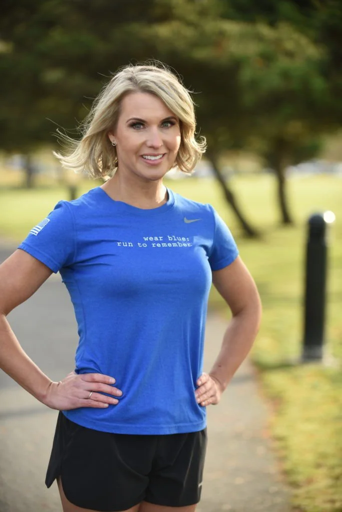 Woman in blue running shirt and black shorts standing outdoors with hands on hips, smiling, with trees in the background.