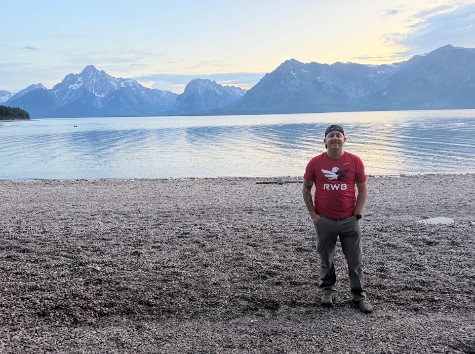 A man wearing a red Nike t-shirt and gray pants stands on a rocky beach by a calm lake with mountain peaks in the background during sunset.