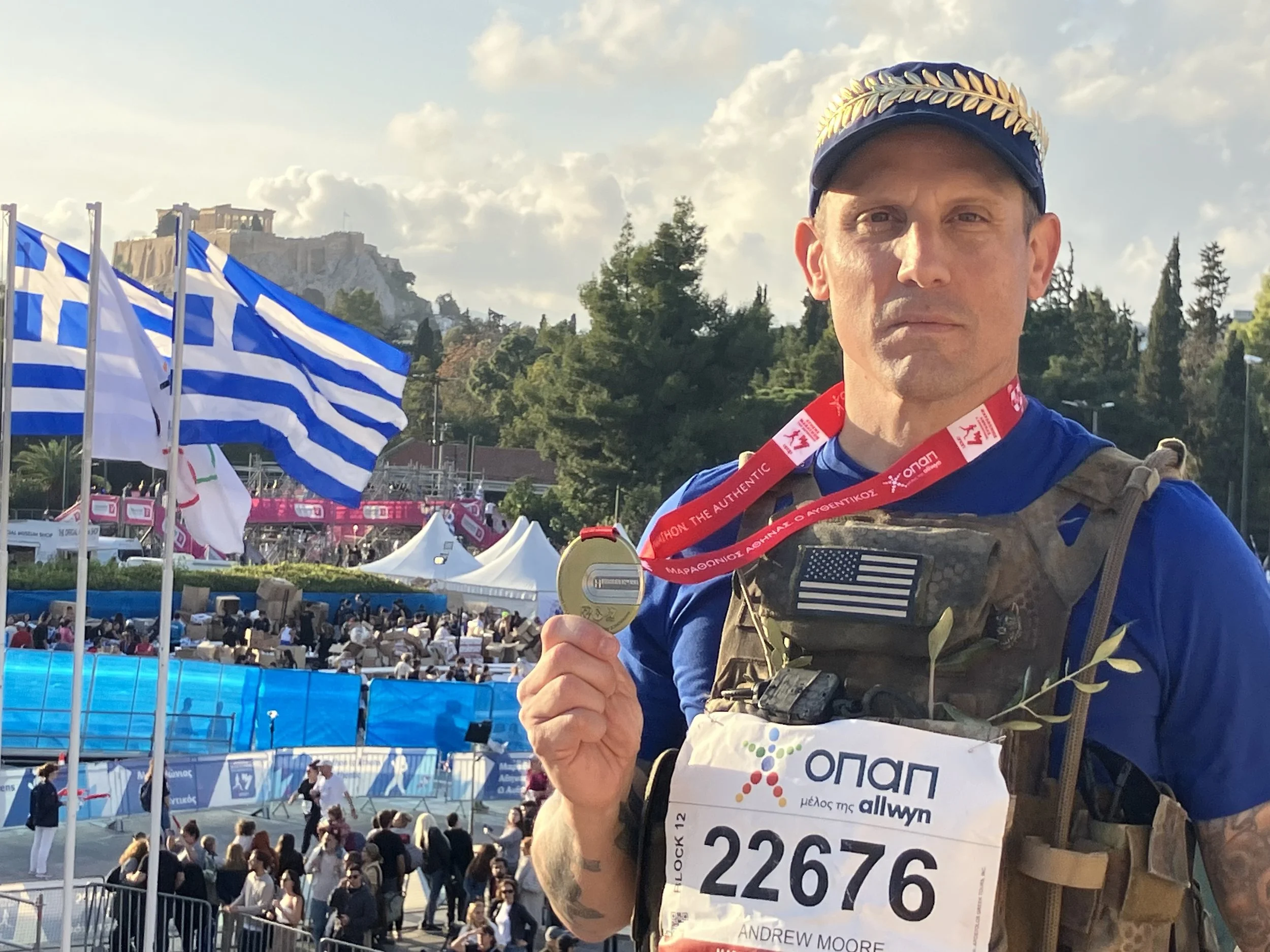 A man with a medal around his neck taking a selfie at a marathon event, with Greek flags, a crowd, and the Acropolis in the background.