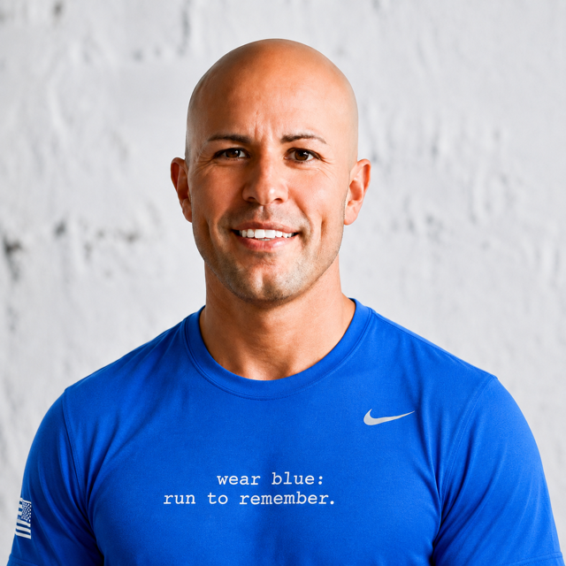 A smiling bald man wearing a blue athletic shirt with the Nike logo and a slogan that reads "wear blue: run to remember" standing in front of a white brick wall.