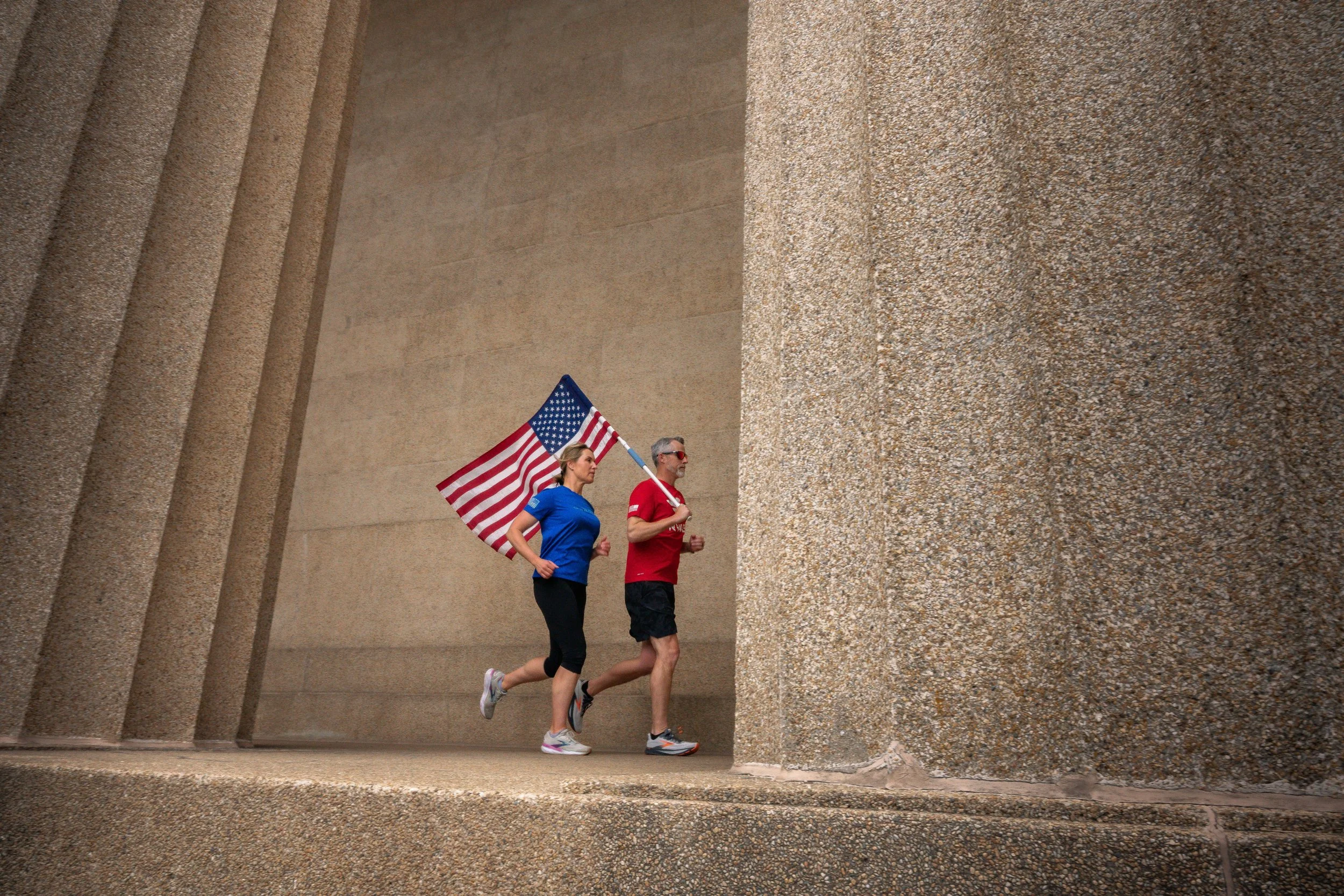 Two people running, one woman and one man, holding American flag, running past large textured concrete walls.