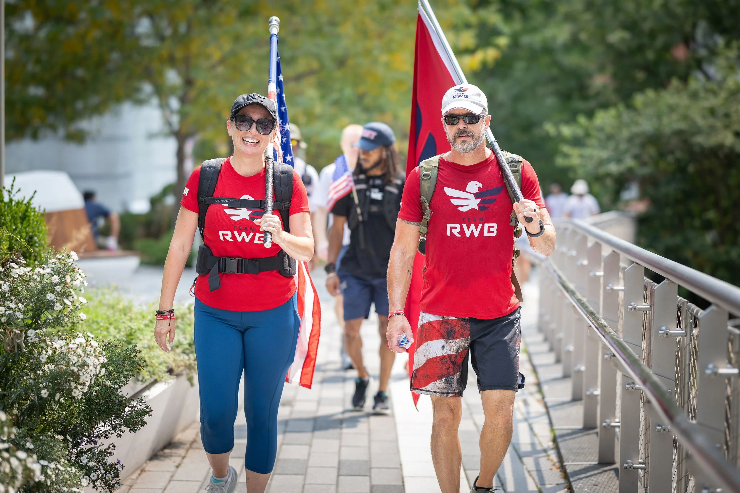 Two people walking outdoors with flags, one woman and one man, both wearing red shirts and backpacks.