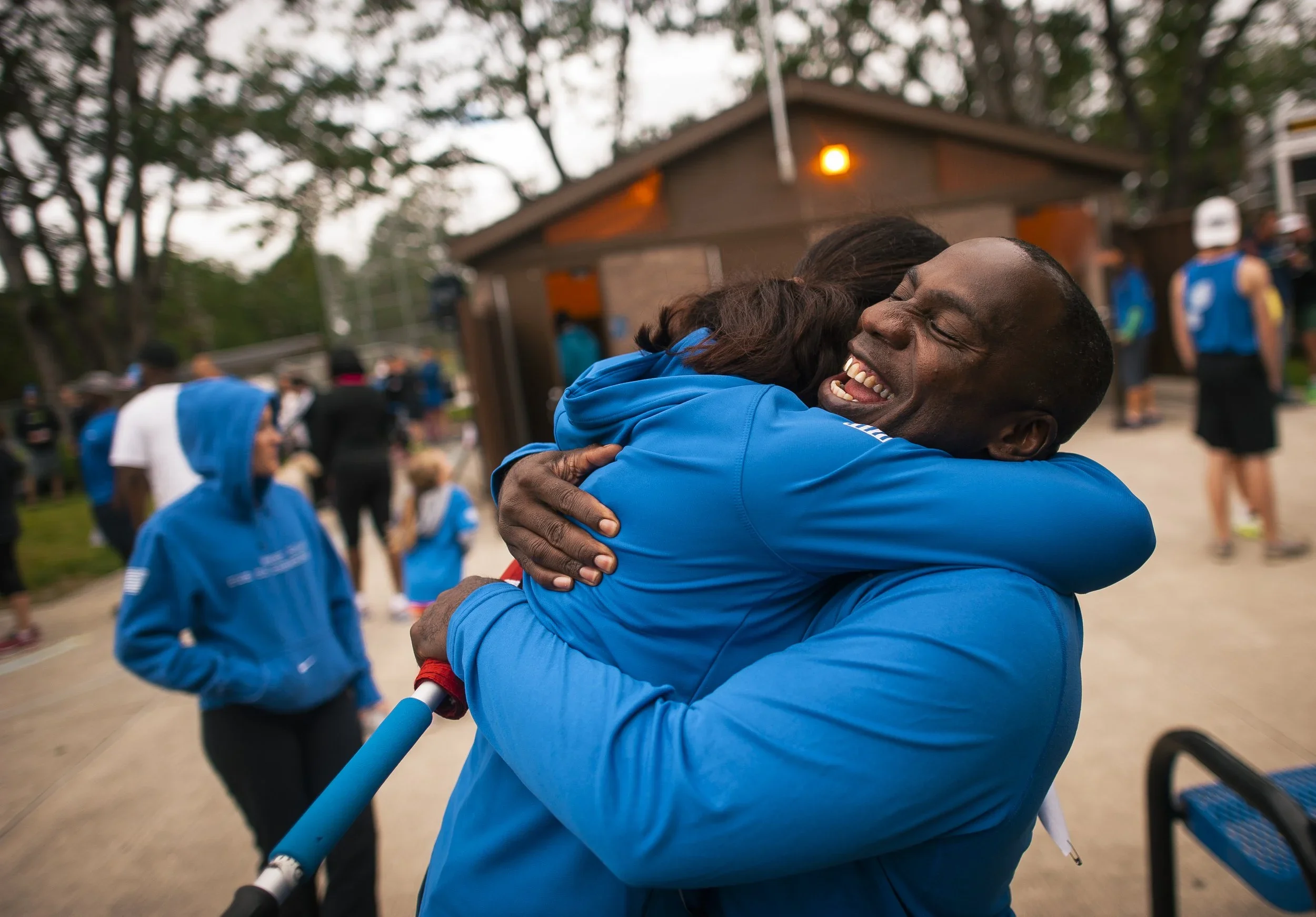 Two people hugging at an outdoor event, with other people in the background, some wearing blue jackets and a building with a warm light above.