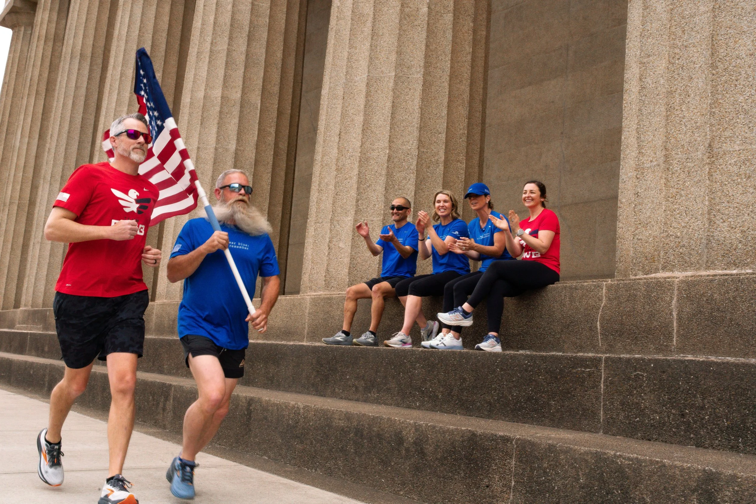 A group of people participating in a race or marathon, with some runners in the foreground and a group of people sitting on steps in the background, clapping and cheering.