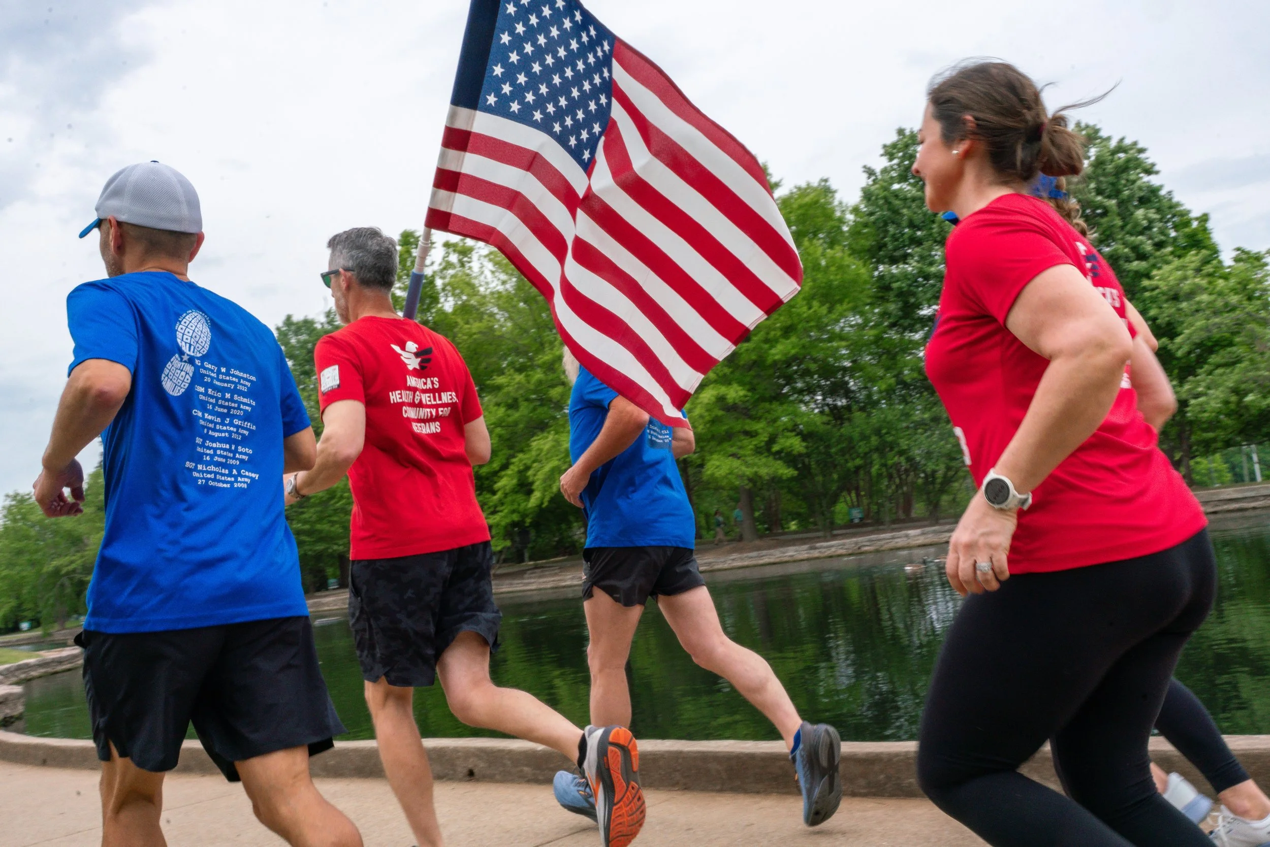 A group of people running outdoors near a body of water, with some wearing shirts that show patriotic and health awareness themes, and an American flag.